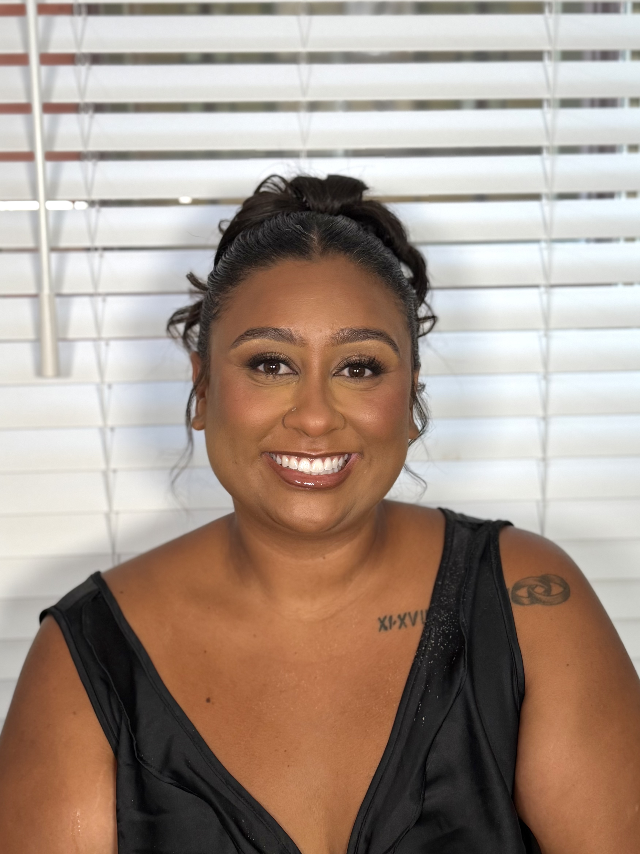 Close-up of a smiling woman with dark hair, tattoos, and a nose piercing, wearing a black sleeveless top, in front of white blinds.