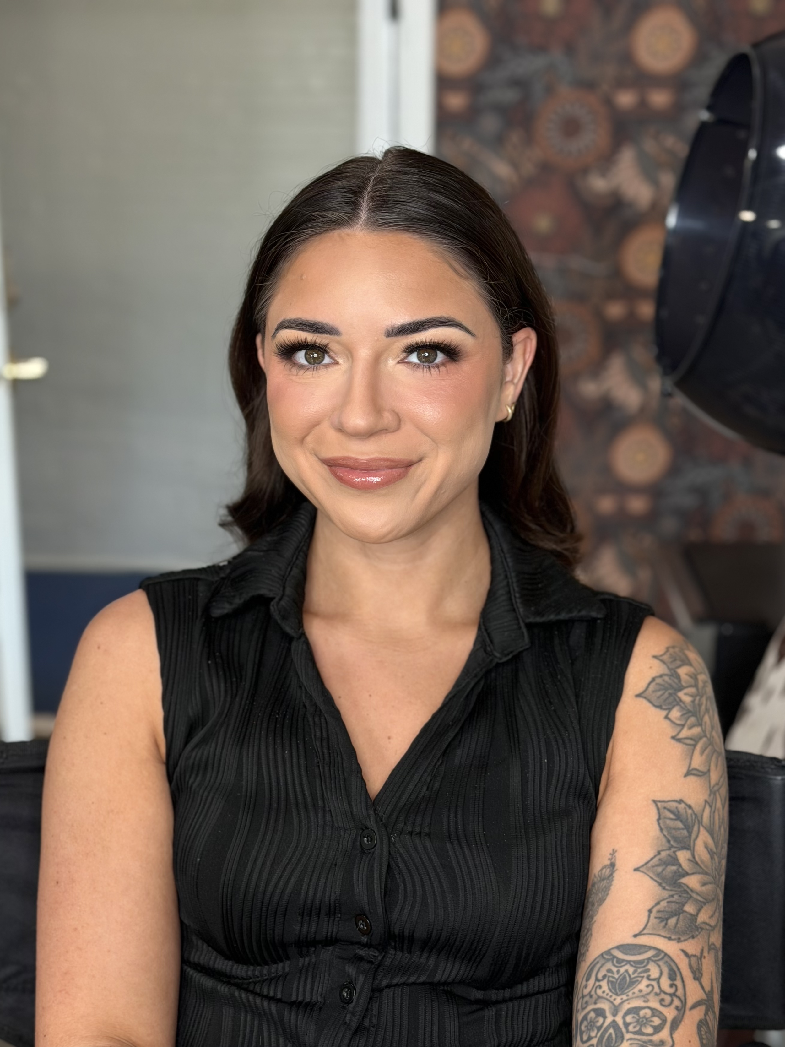 A woman with long brown hair and tattoos on her left arm, wearing a sleeveless black button-up top, smiling in a room with patterned flooring and a speaker in the background.