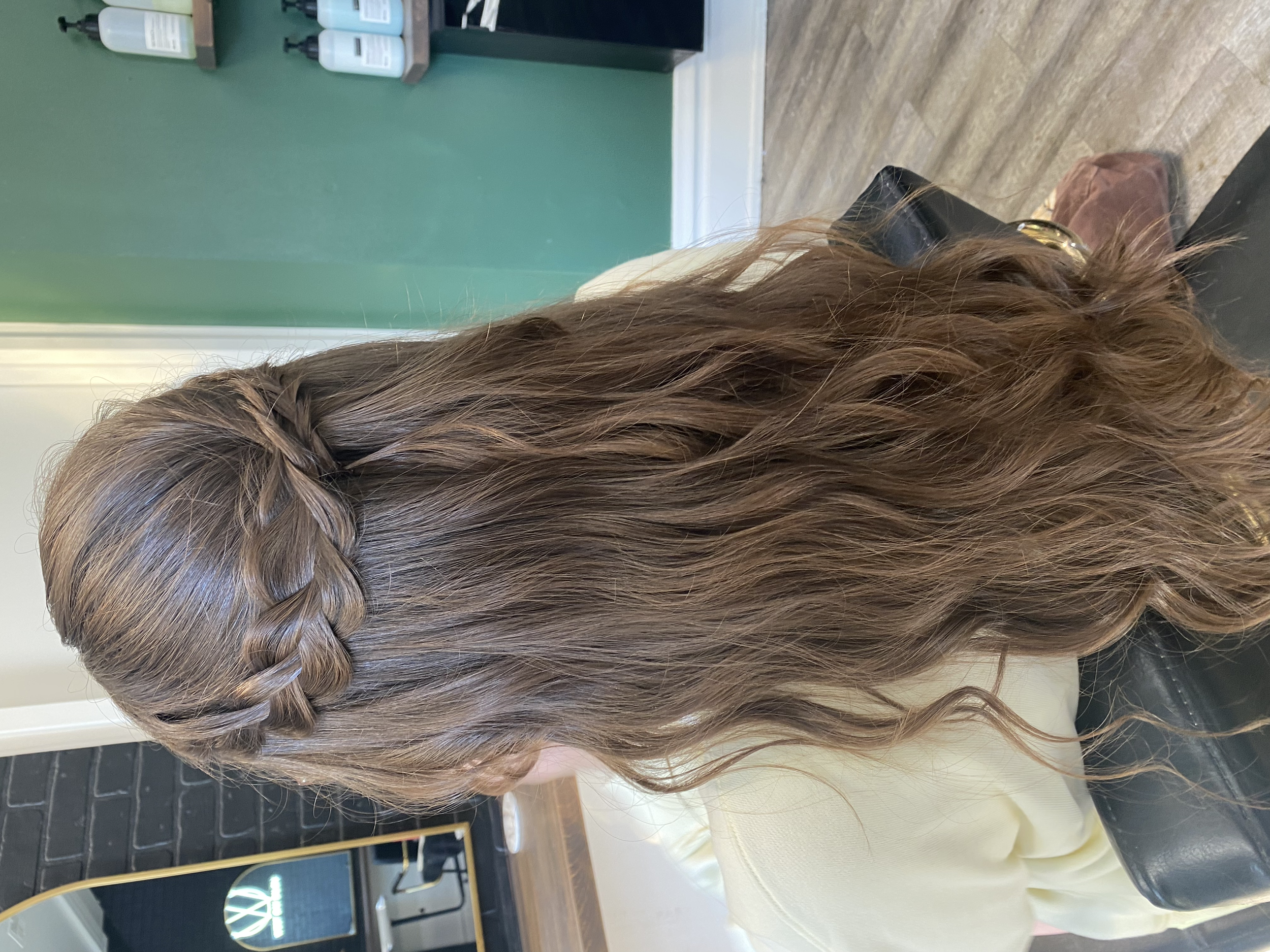 A person with long, wavy, light brown hair sitting in a black salon chair at a hair salon, with their head slightly turned.