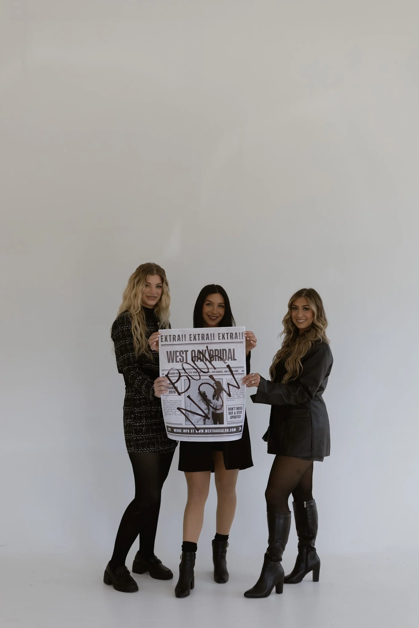 Three women standing together, smiling, holding a large printed advertisement for a bridal event, with a plain white background.