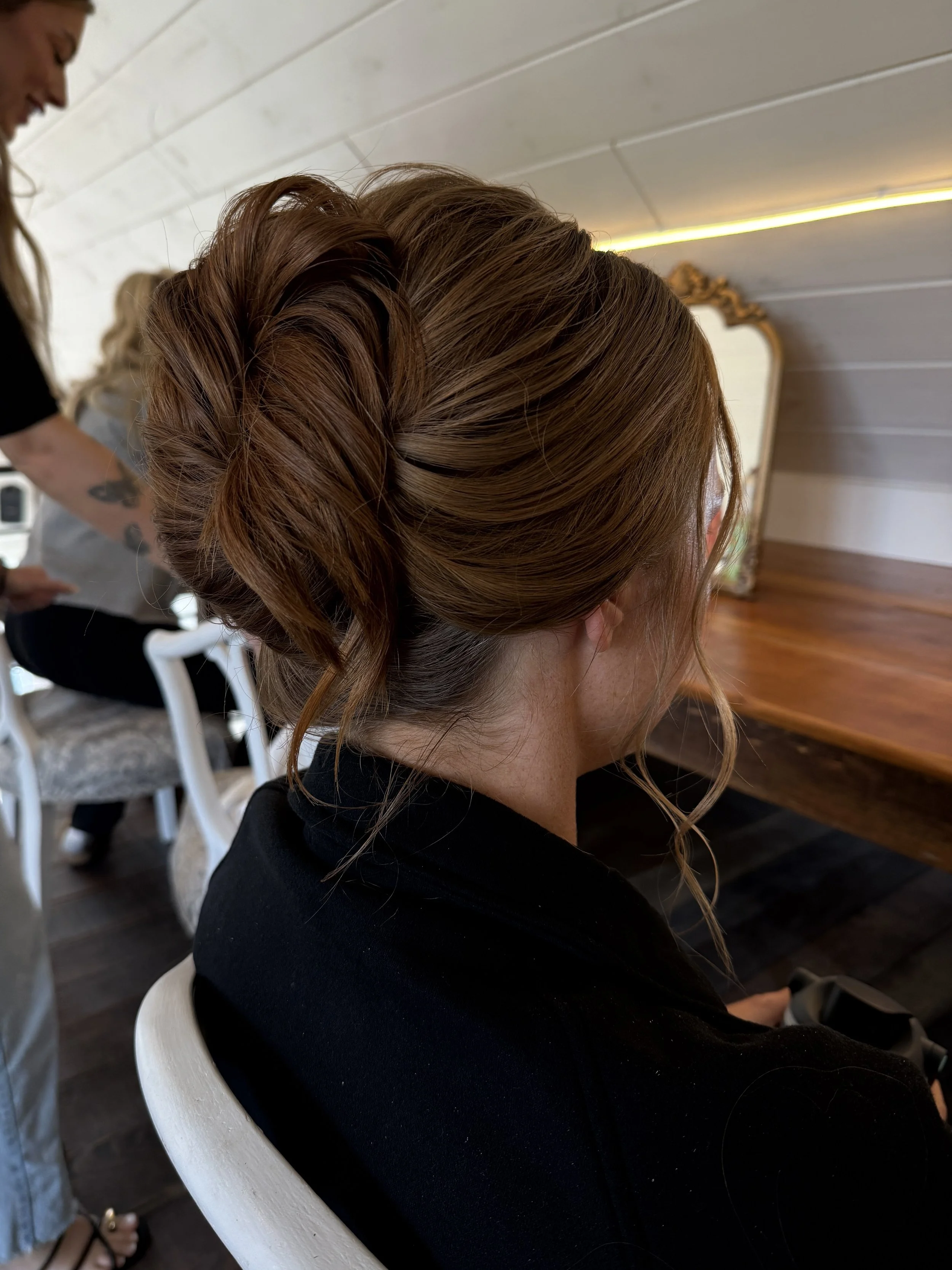Side view of a woman with styled auburn hair sitting in a chair, with a mirror and a hairstylist visible in the background.