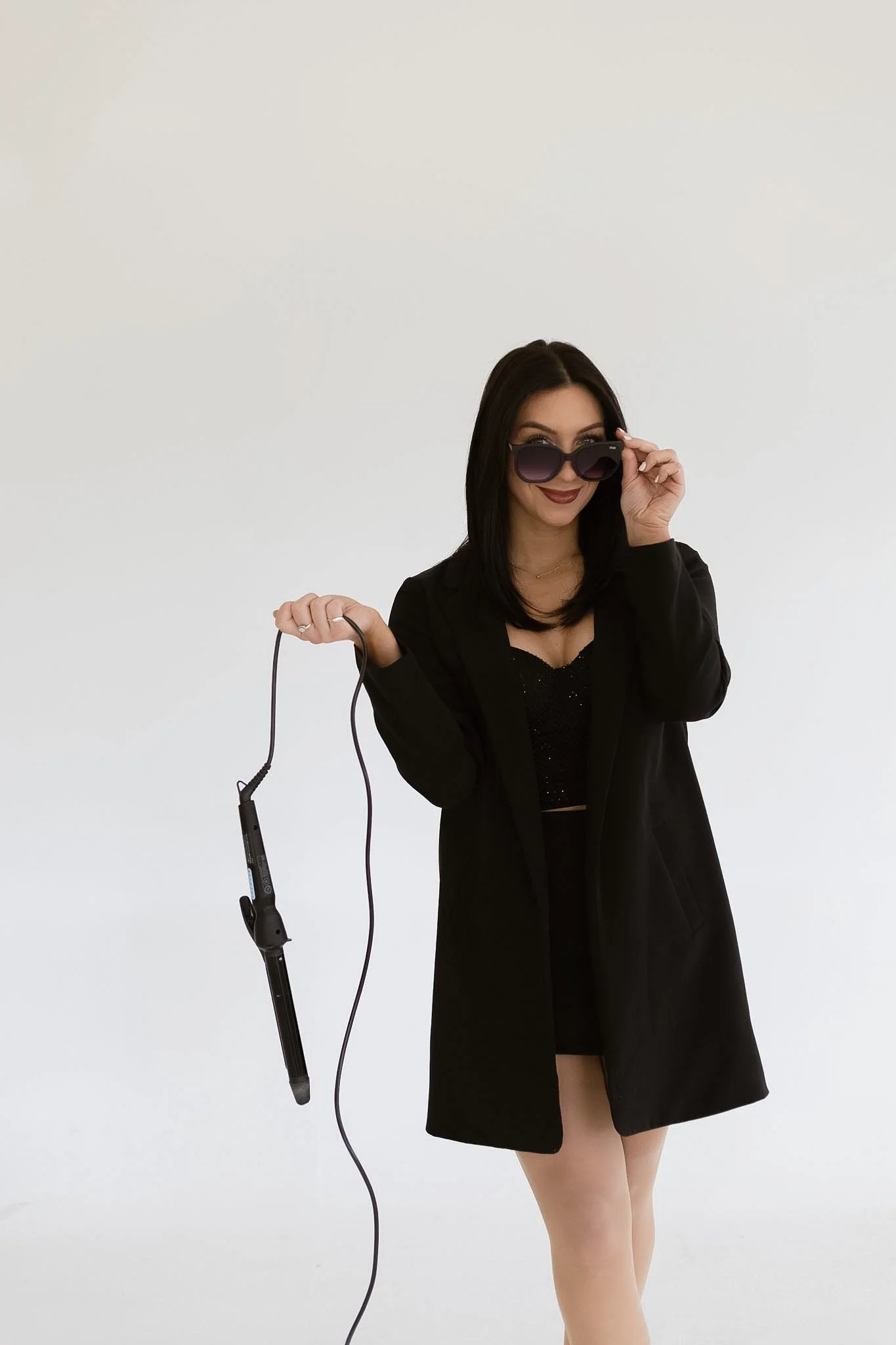 A woman with black hair wearing sunglasses, a black blazer, and a black dress, holding a hair straightener and smiling.