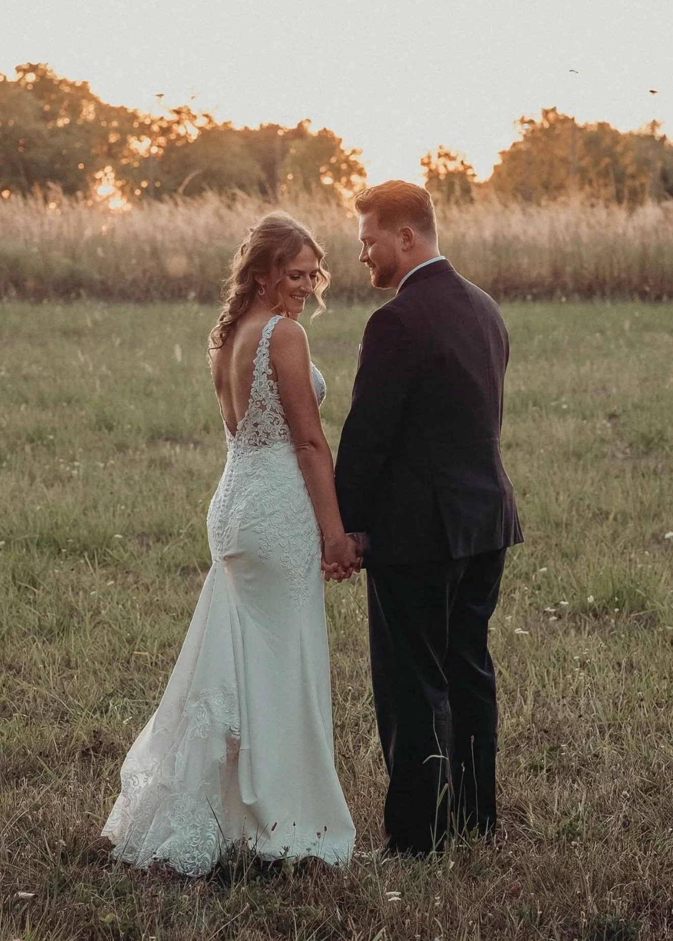 A bride and groom holding hands and smiling at each other in an open field during sunset, dressed in wedding attire.