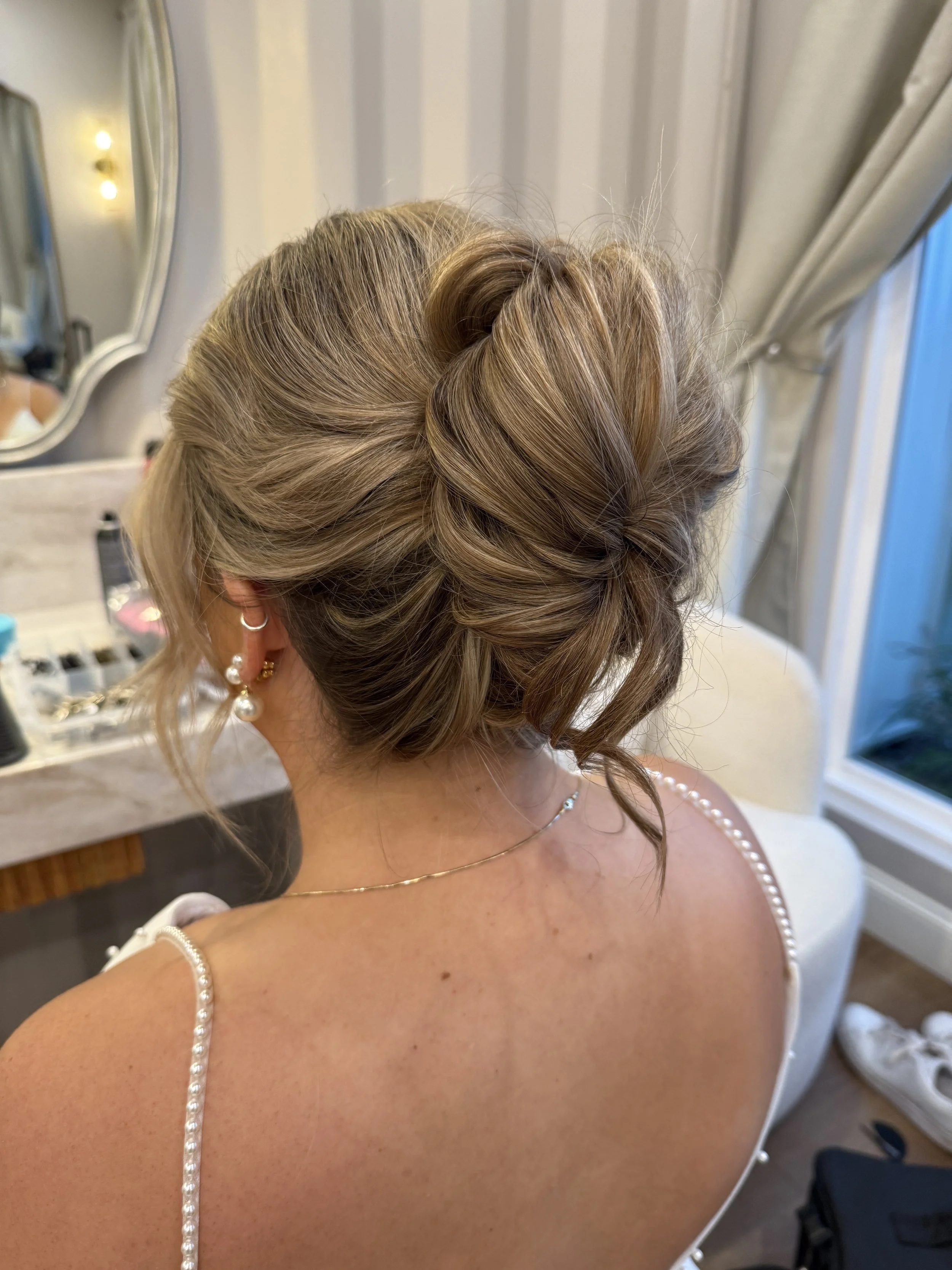 A woman with styled blonde hair wearing pearl earrings and a pearl strap dress, seen from the back in a room with a mirror and makeup items.