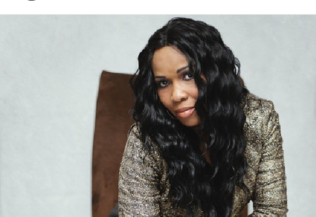 A woman with long, curly black hair wearing a patterned gold and black blazer sitting in a chair against a plain light-colored wall.