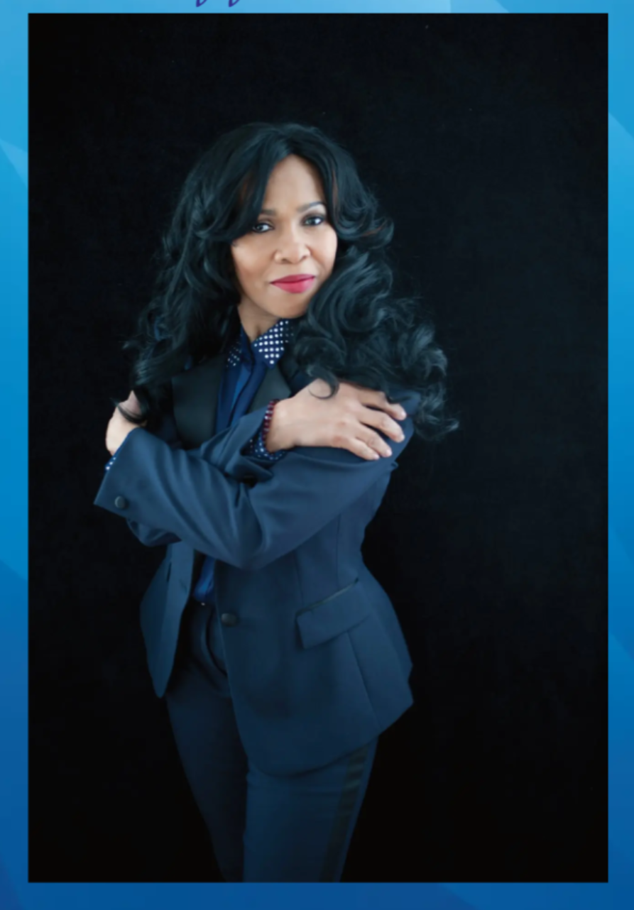 A woman with long black curly hair wearing a dark blue suit with a polka dot shirt, standing against a black background, crossing her arms and looking confidently at the camera.