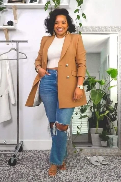 A smiling woman with curly black hair wearing a tan blazer, white top, and ripped jeans, standing in a room with plants and a clothing rack.