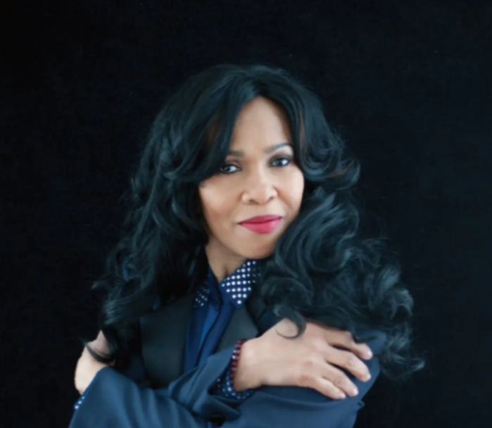 A woman with black, curly hair and red lipstick wearing a dark blazer and polka dot shirt, standing against a black background.