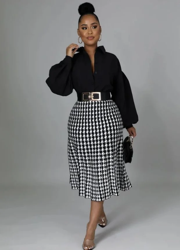 Woman with dark hair in a bun, wearing a black blouse with balloon sleeves, a black and white houndstooth skirt, and clear high heels. She's accessorized with large hoop earrings and a black handbag, standing against a plain gray background.