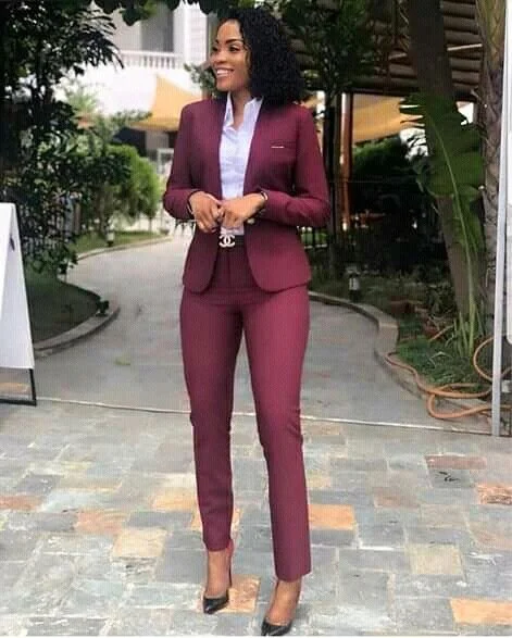 A woman in a burgundy business suit with a white blouse, standing outdoors on a paved pathway surrounded by greenery, smiling and looking to her left.