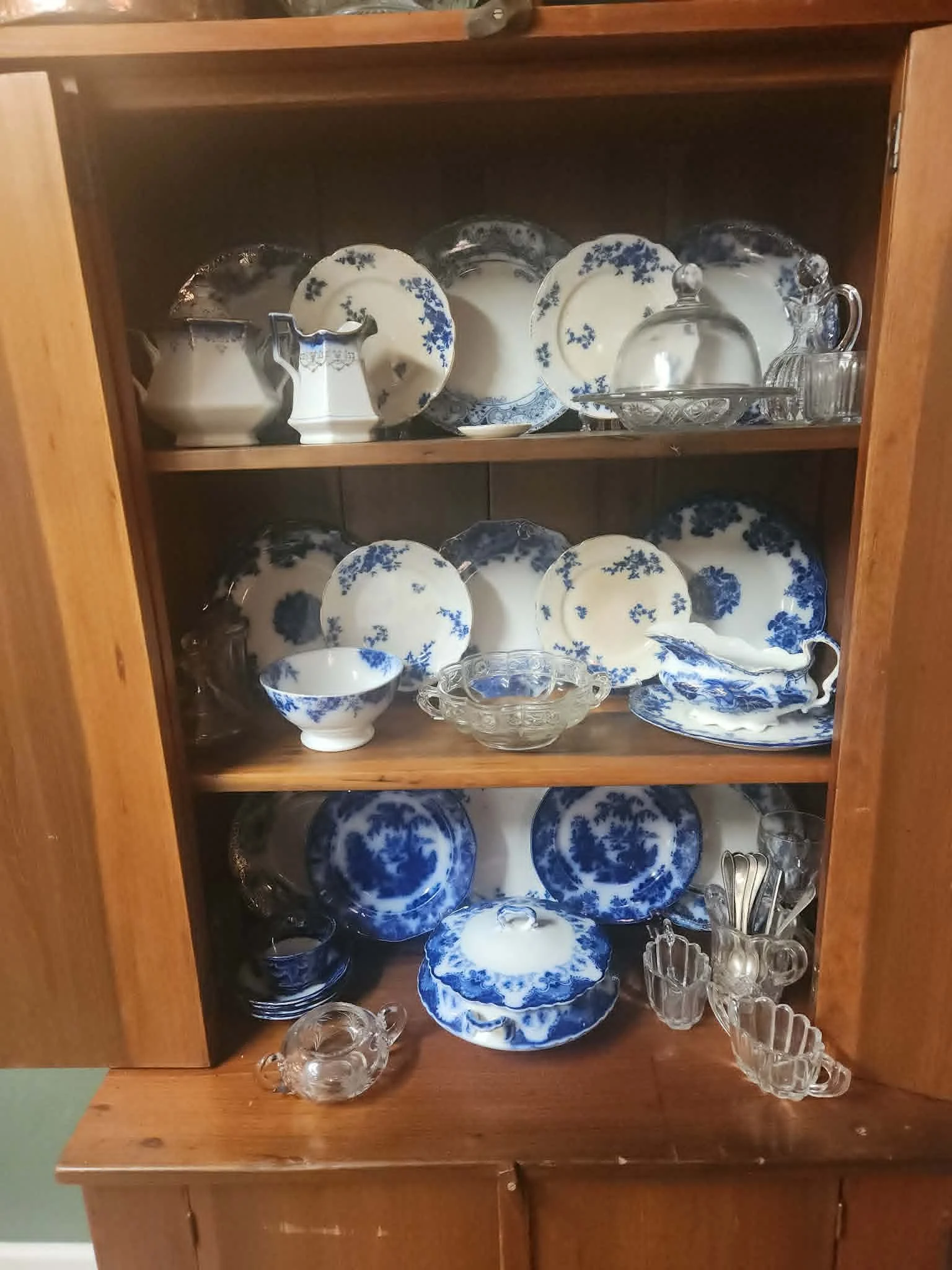 A wooden cabinet displaying blue and white china plates, bowls, and teapots, along with clear glass cups and serving dishes.
