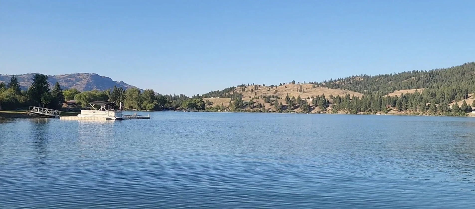 A calm lake with a dock and boat in the foreground, surrounded by trees and rolling hills in the distance under a clear blue sky.