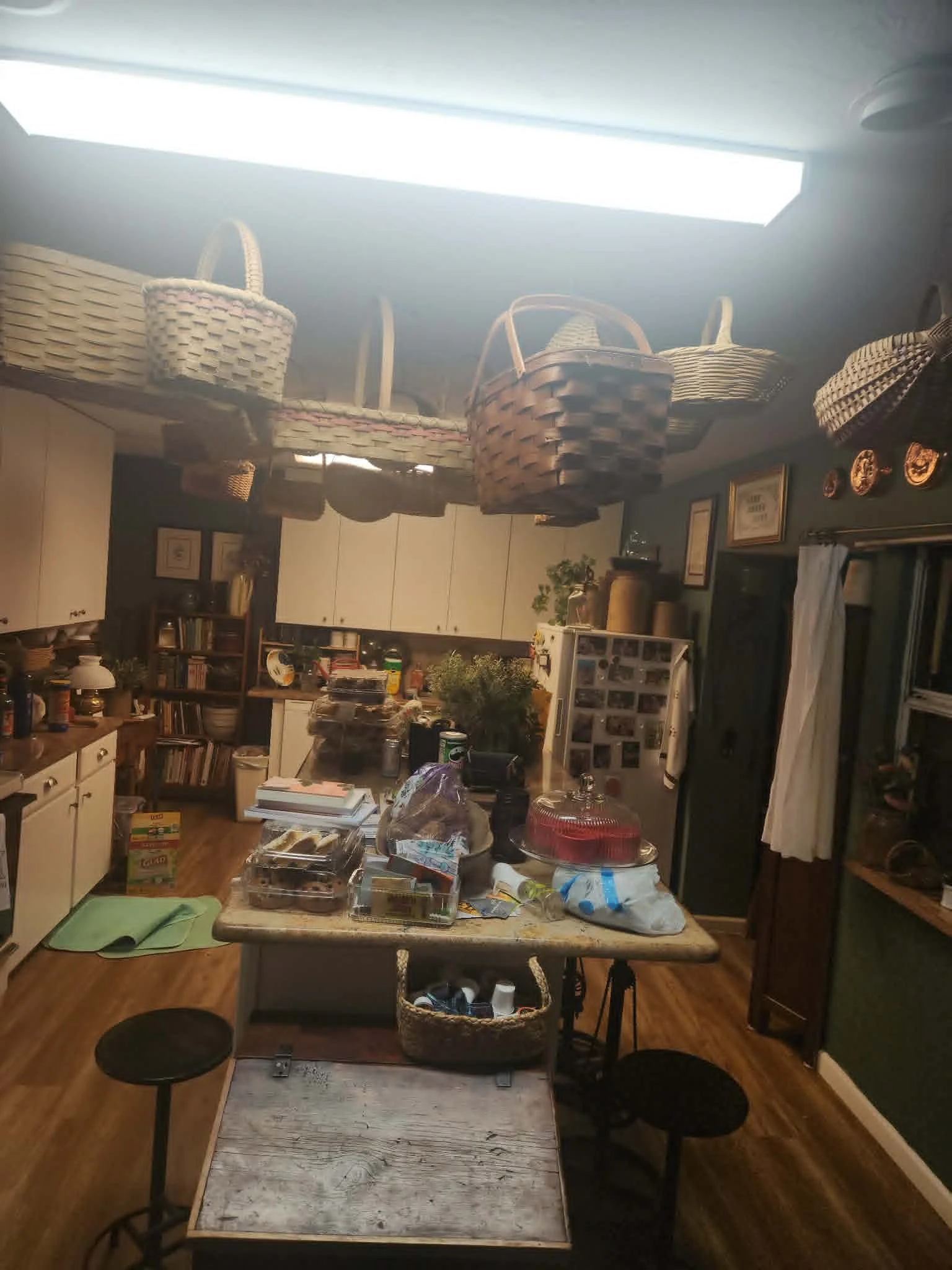 A kitchen with a kitchen table covered in various items including food containers, books, and bags. The room has white cabinets, a bookshelf, and hanging baskets on the ceiling. There is a window with curtains and framed artwork on the walls.