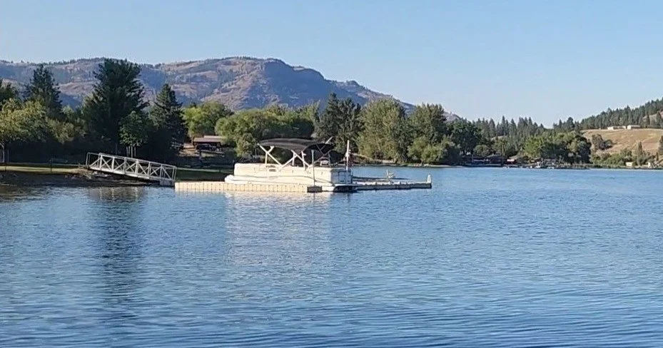 Calm lake with a docked boat, surrounded by trees and hills in the distance under a clear sky.