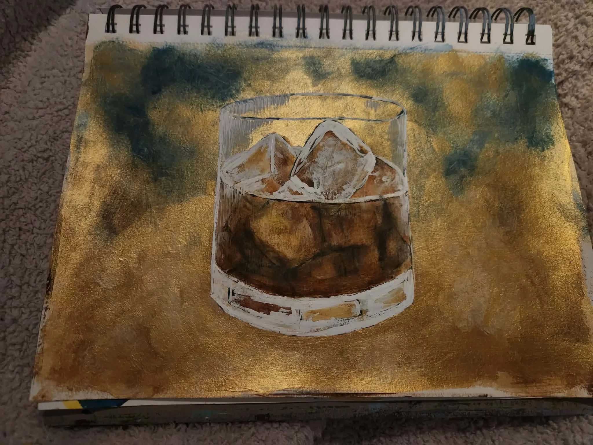 Original painting. A spiral-bound sketchbook on a carpeted surface, featuring an abstract painting of a glass with ice cubes, painted with gold, brown, and white tones.