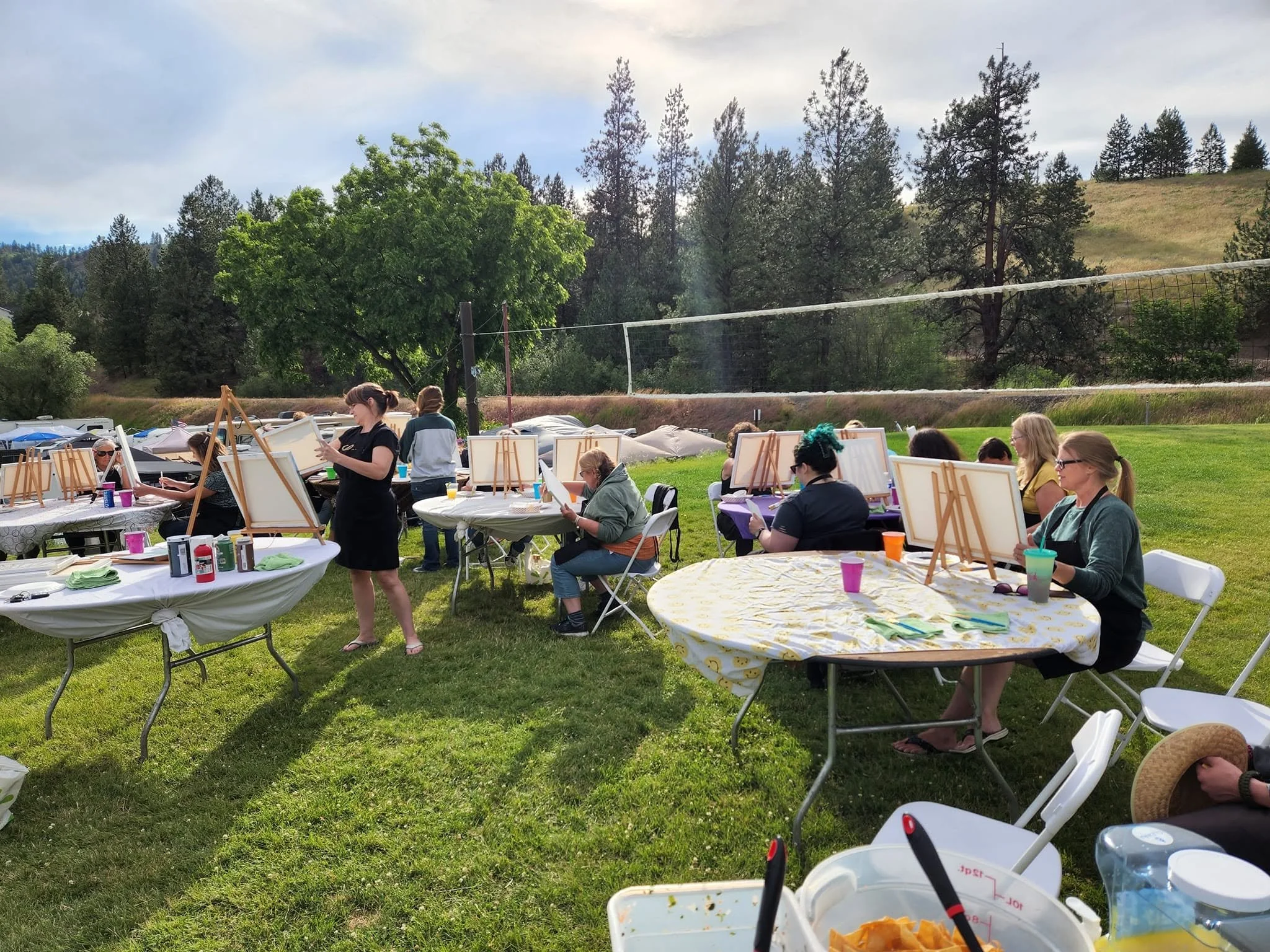 People participating in outdoor painting activity in a park, with easels and art supplies on tables, surrounded by trees and grassy hills.