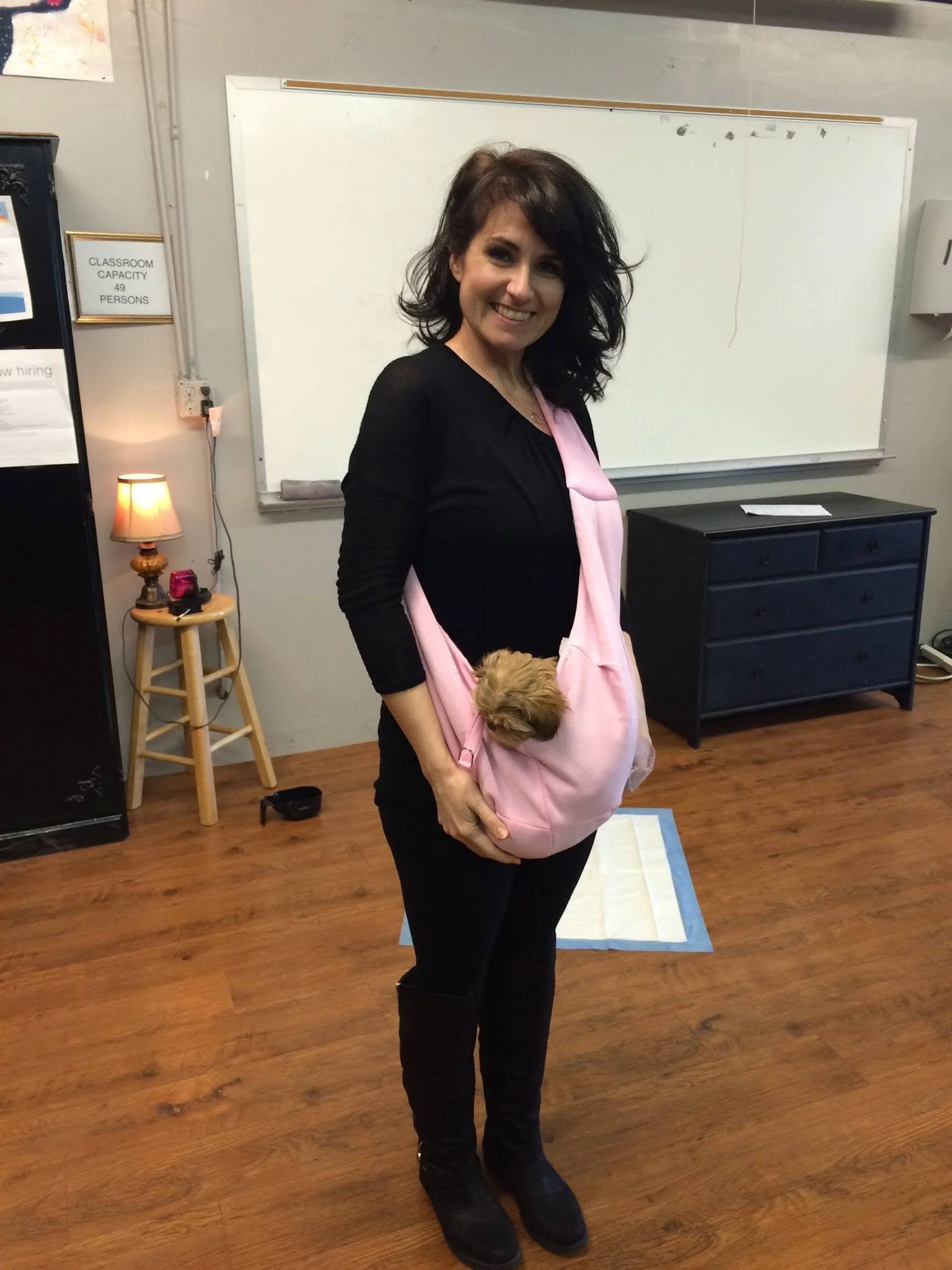 Woman smiling while standing in a room, holding a pink baby carrier with a small brown puppy inside. The room has a whiteboard, a small dresser, and a lamp on a stool.