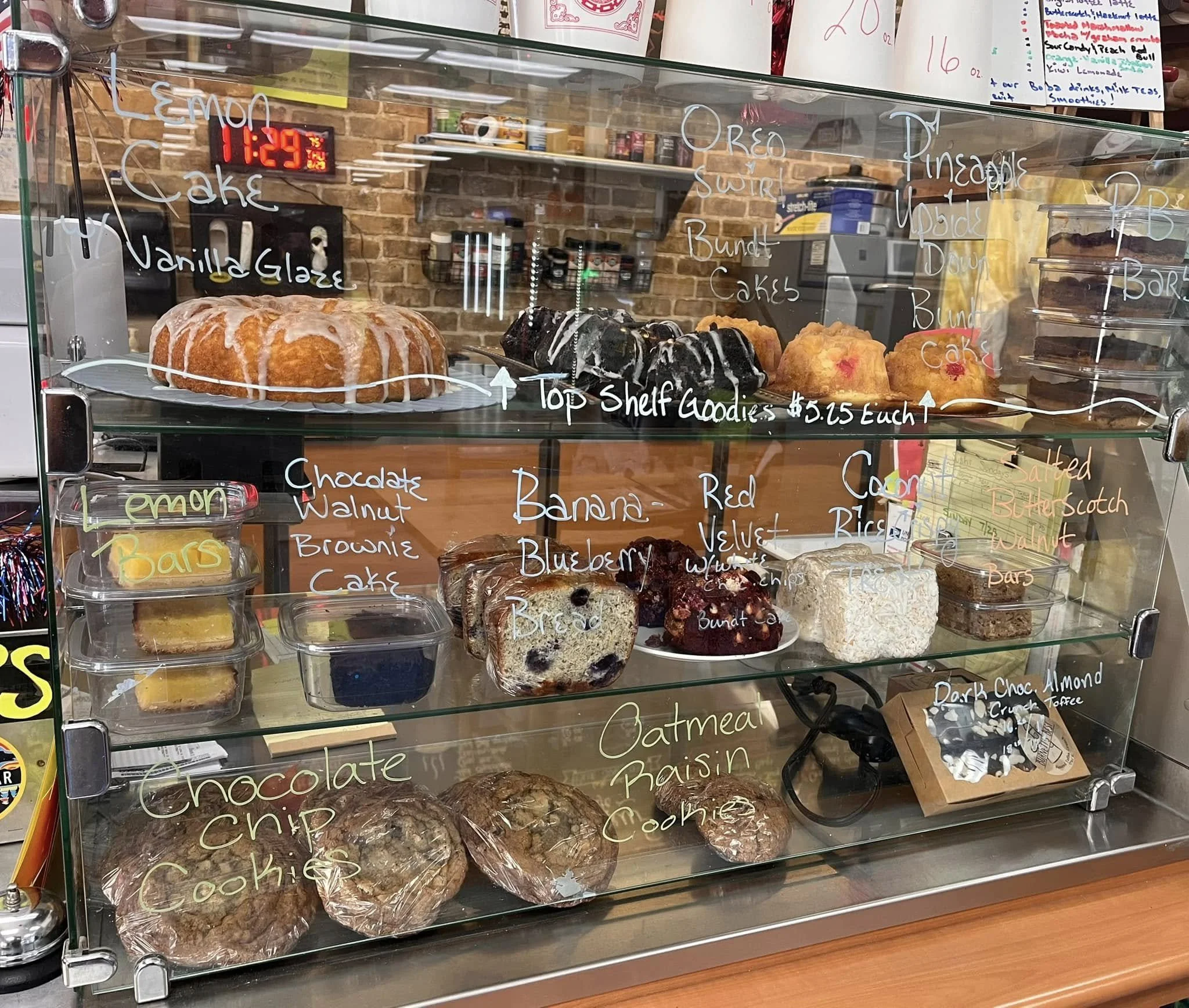 Display case with various baked goods including lemon cake, chocolate chip cookies, blueberry muffins, banana bread, and oatmeal raisin cookies, with handwritten labels and prices on the glass.