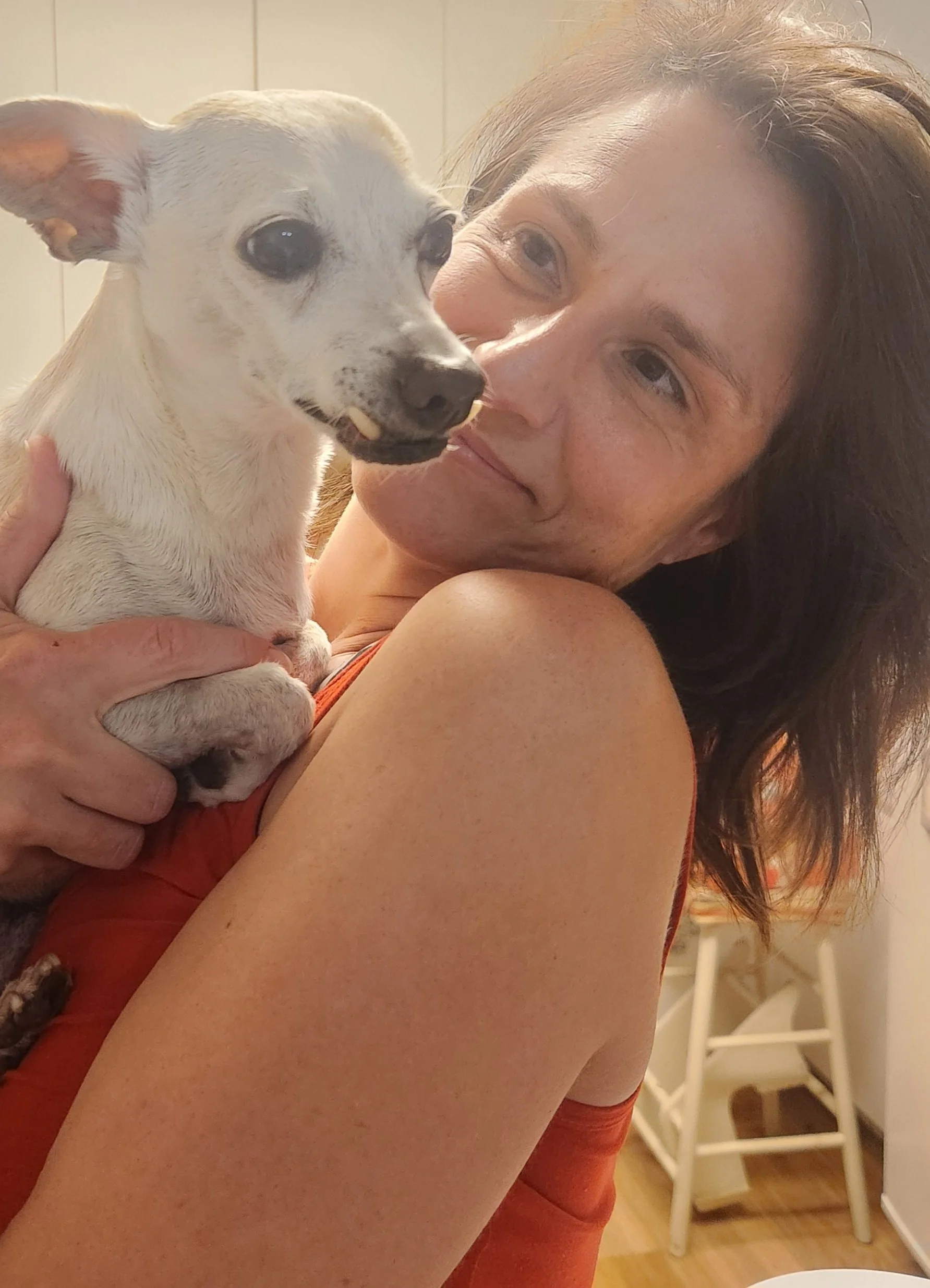A woman holding a small white dog with black spots close to her face, both looking at the camera, indoors with a wooden floor and white chairs in the background.