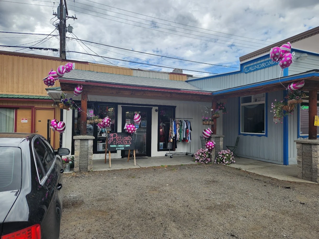Storefront decorated with pink polka dot and striped balloons, flowers, and clothing outside Petticoats & Pistols, with a parking lot in front and a cloudy sky above.