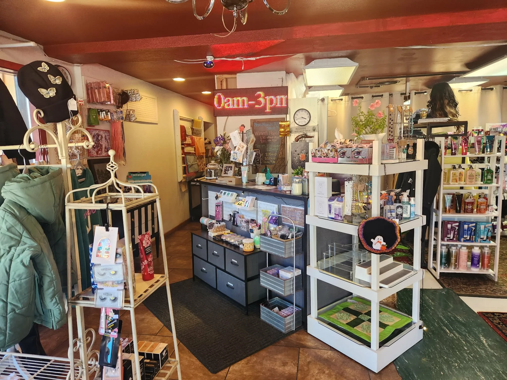The interior of a small retail store with various display racks and shelves filled with hair and beauty products. There is a blackboard with handwritten prices, a red LED sign indicating the store's hours from 9 am to 3 pm, and a woman with curly dark hair looking at products on the right side. The ceiling has a chandelier and a security camera.