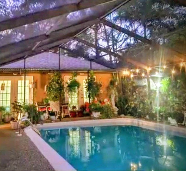 Outdoor swimming pool surrounded by tropical plants and a screened enclosure with warm evening lights.