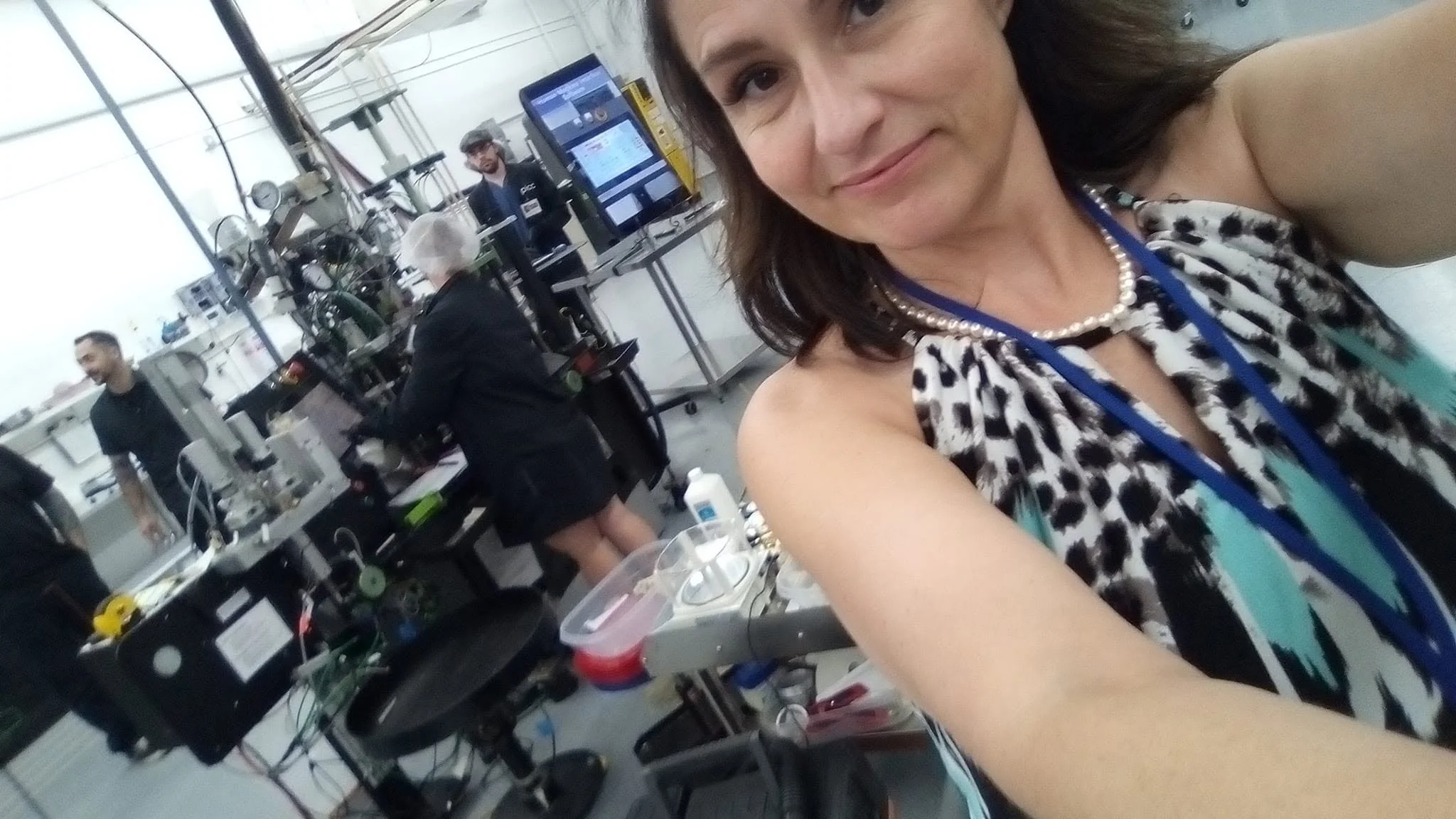 A woman taking a selfie in a laboratory with scientists working in the background surrounded by scientific equipment and computers.
