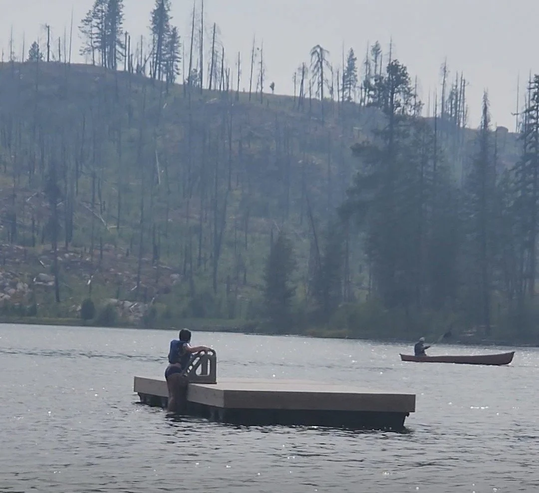 A person standing on a floating dock and fishing in a lake, with a small boat and another person fishing in the background, surrounded by trees and hills.