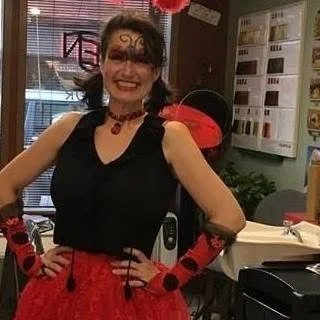 A woman dressed in Gothic style with black and red clothing, face makeup, and gloves, smiling indoors.