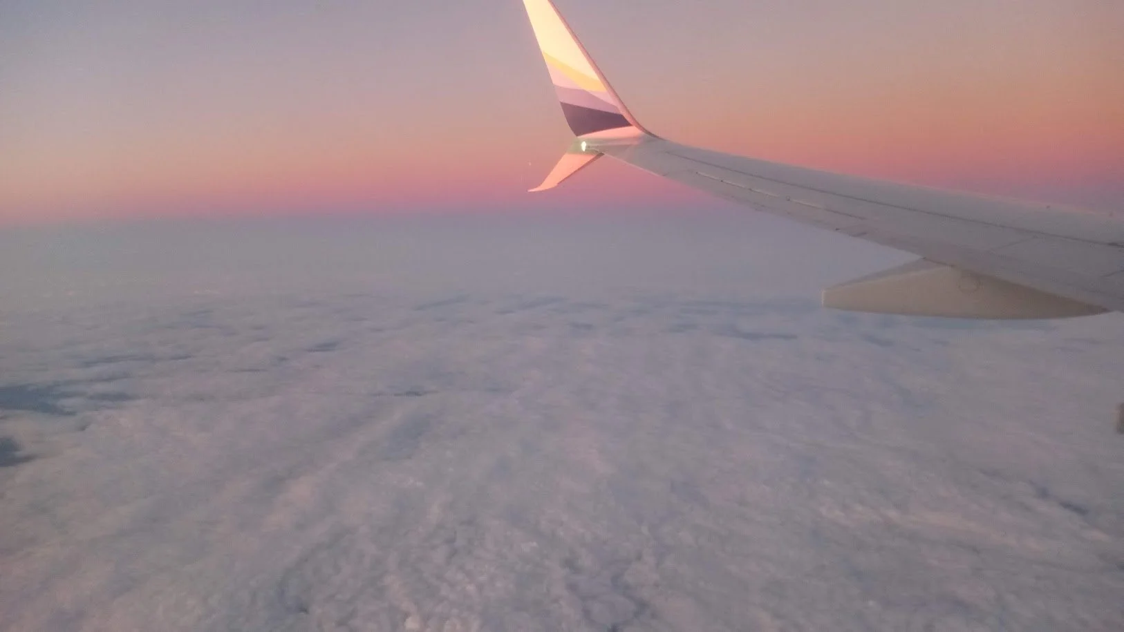 View from an airplane window showing the wing and sunrise over snowy clouds.