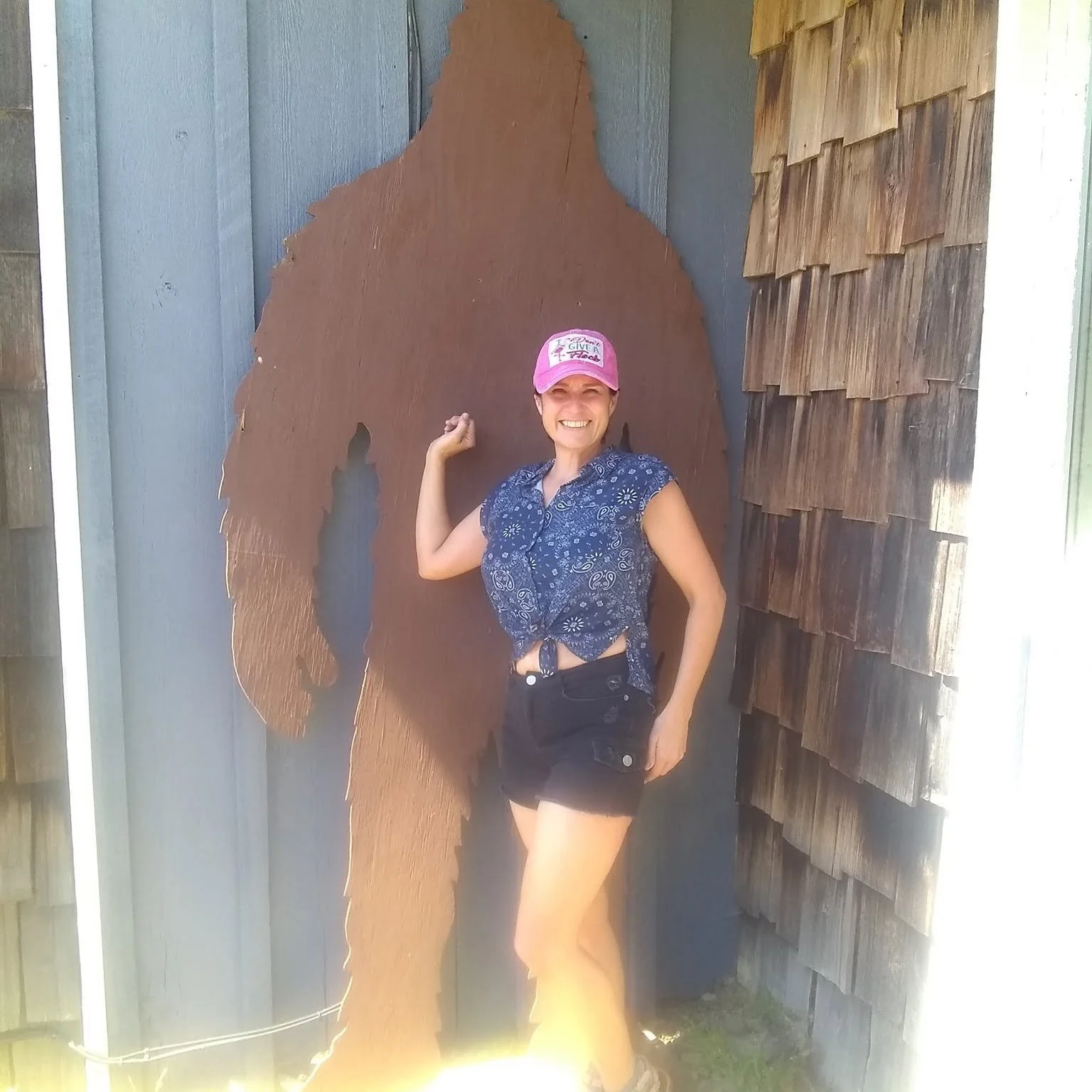A woman smiling and wearing a pink cap with text, a blue patterned sleeveless shirt, and black shorts, standing outdoors in front of a large wooden cutout of a bear.