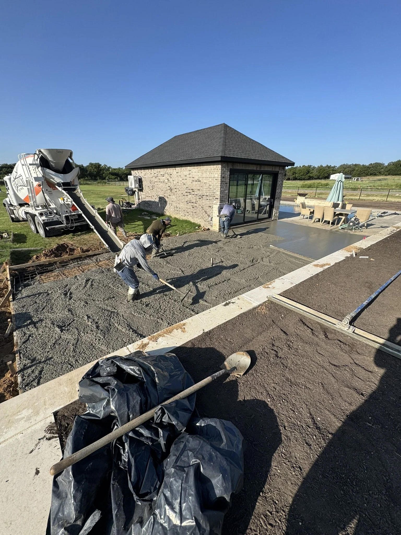 Construction workers are pouring and smoothing concrete in a backyard patio area. There is a small brick building with large glass doors, outdoor furniture, and a concrete mixer truck nearby. The sky is clear and the area is surrounded by green fields.