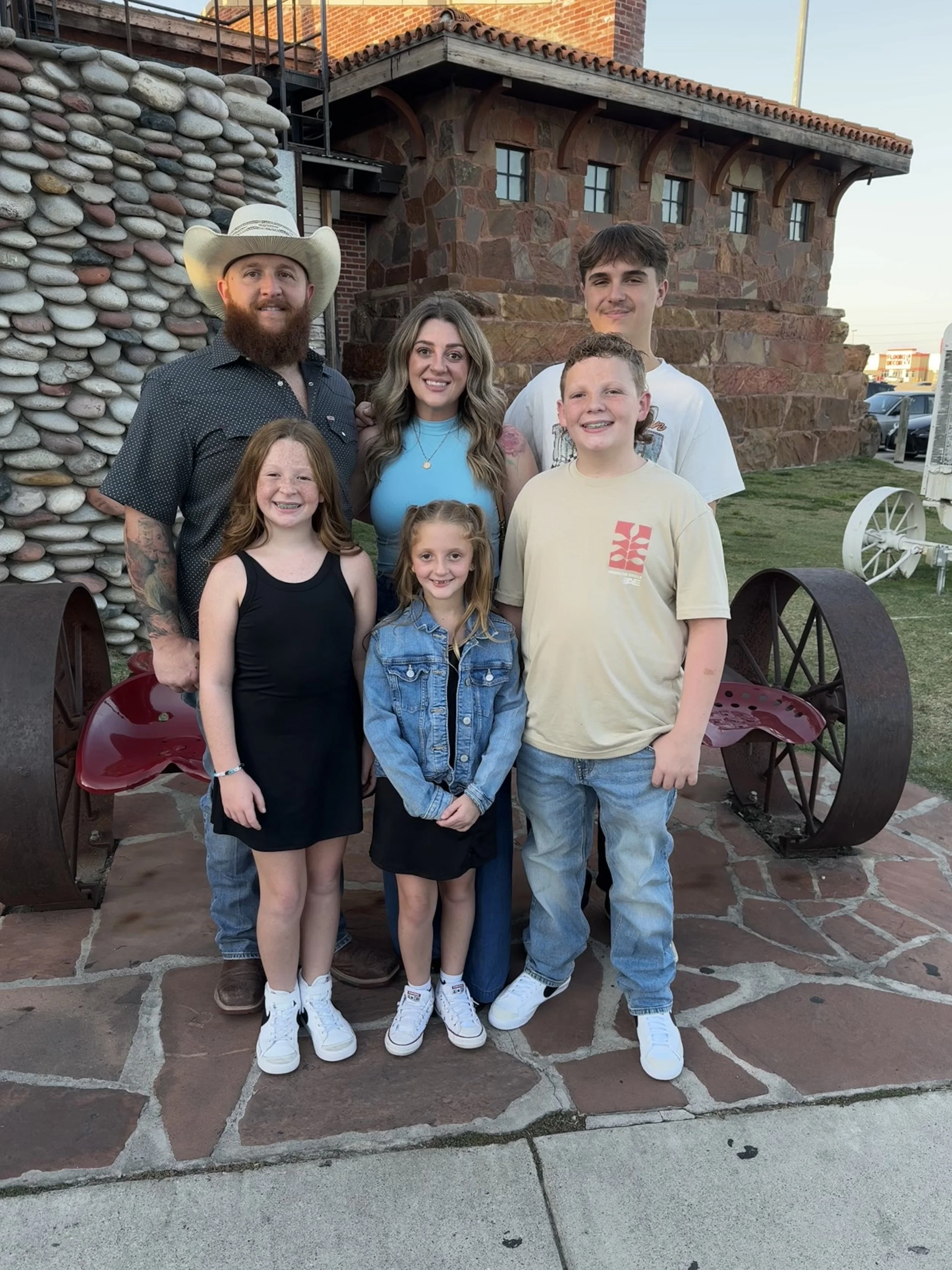 A family of six posing outdoors in front of a building with stone and brick walls. The group includes two young girls, a teenage boy, and three adults. They are smiling and dressed casually, with the man on the left wearing a cowboy hat.