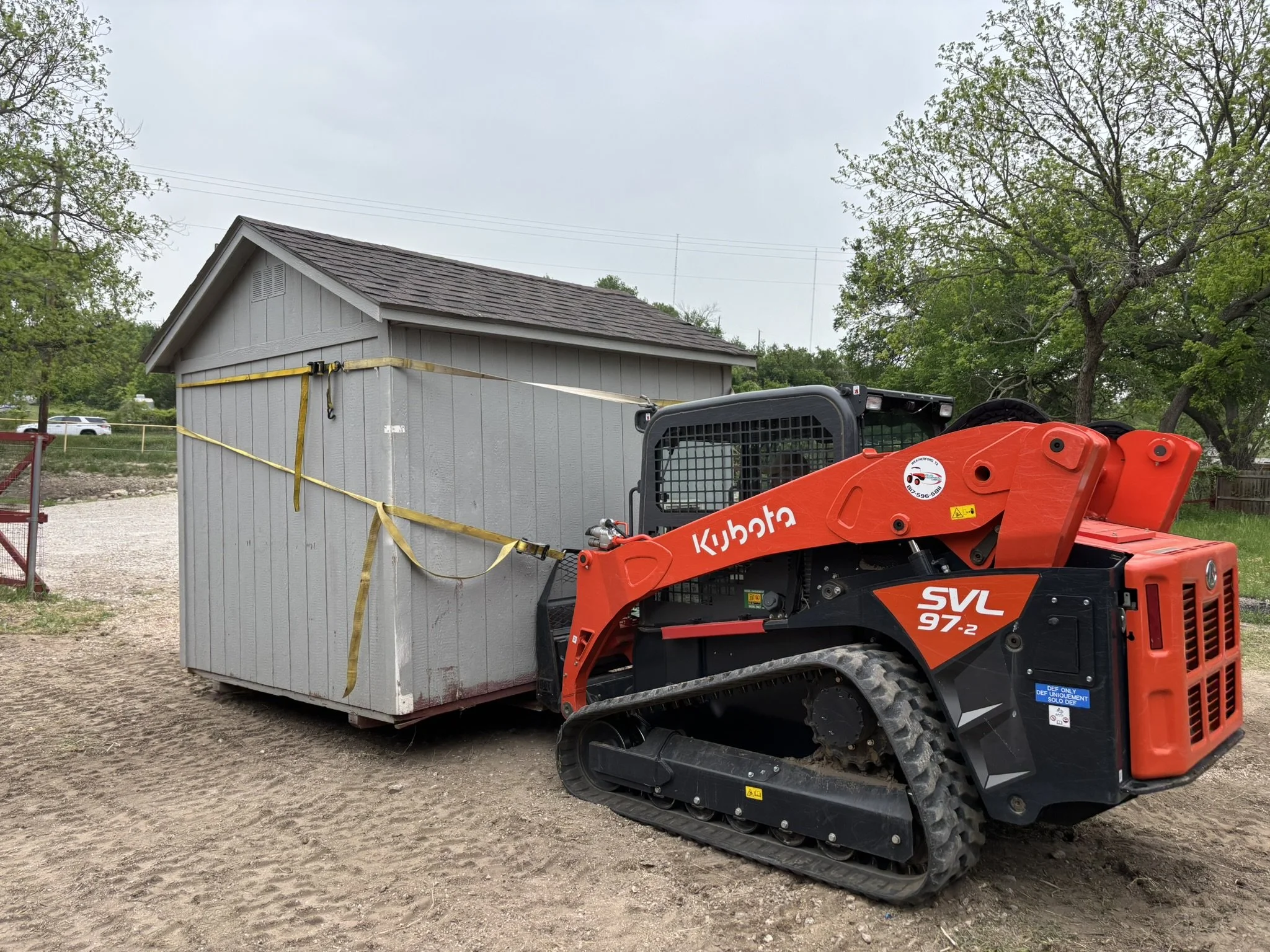 A construction site features a small gray wooden building with a shingle roof. A red Kubota SVL 97-2 compact track loader is nearby, and yellow caution tape is used for safety around the building. Green trees and a gravel ground are in the background.