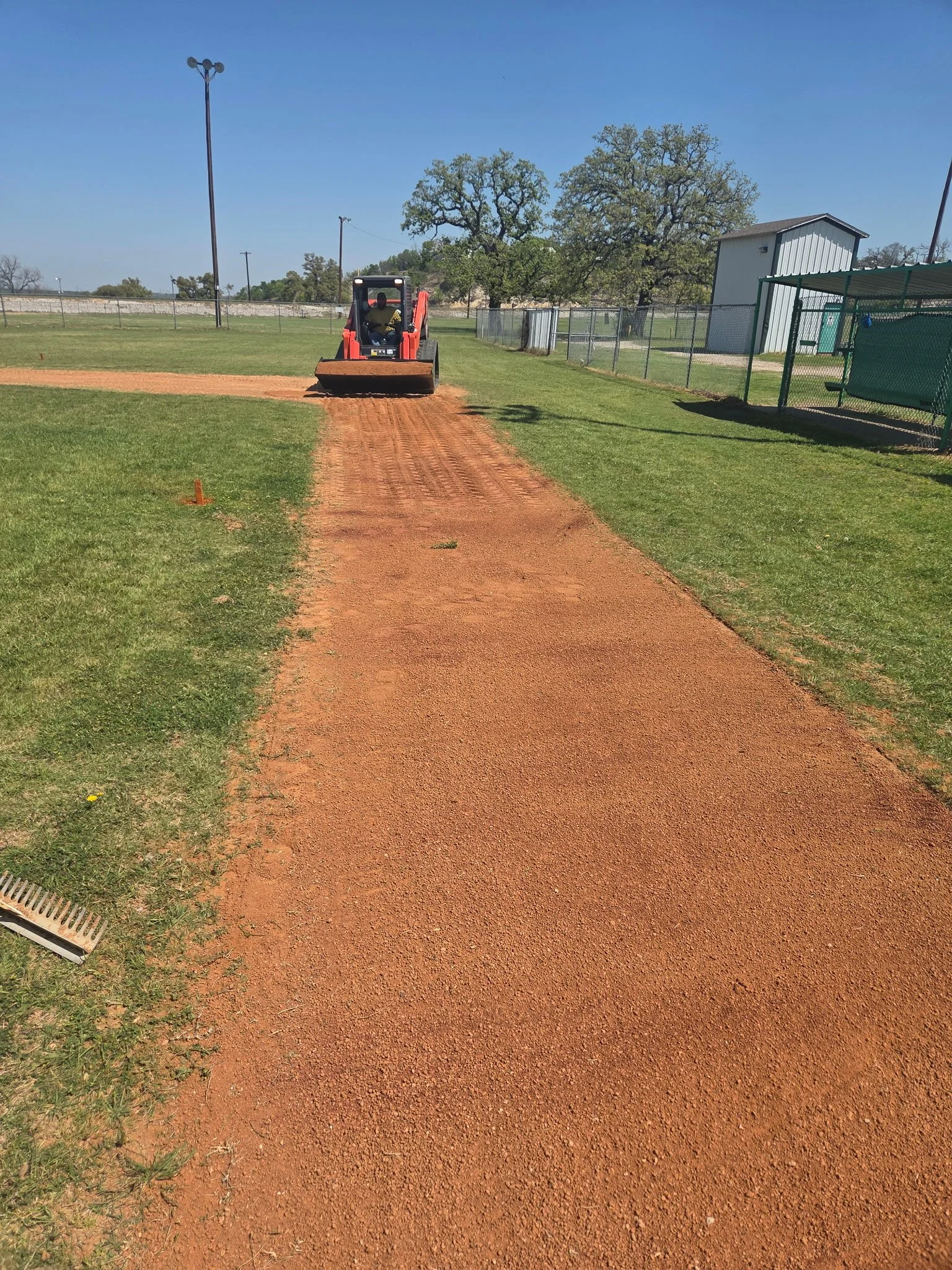 A person operating a small red tractor on a baseball field, leveling the dirt along the base path. The field is surrounded by a chain-link fence, with trees and a building in the background under a clear blue sky.