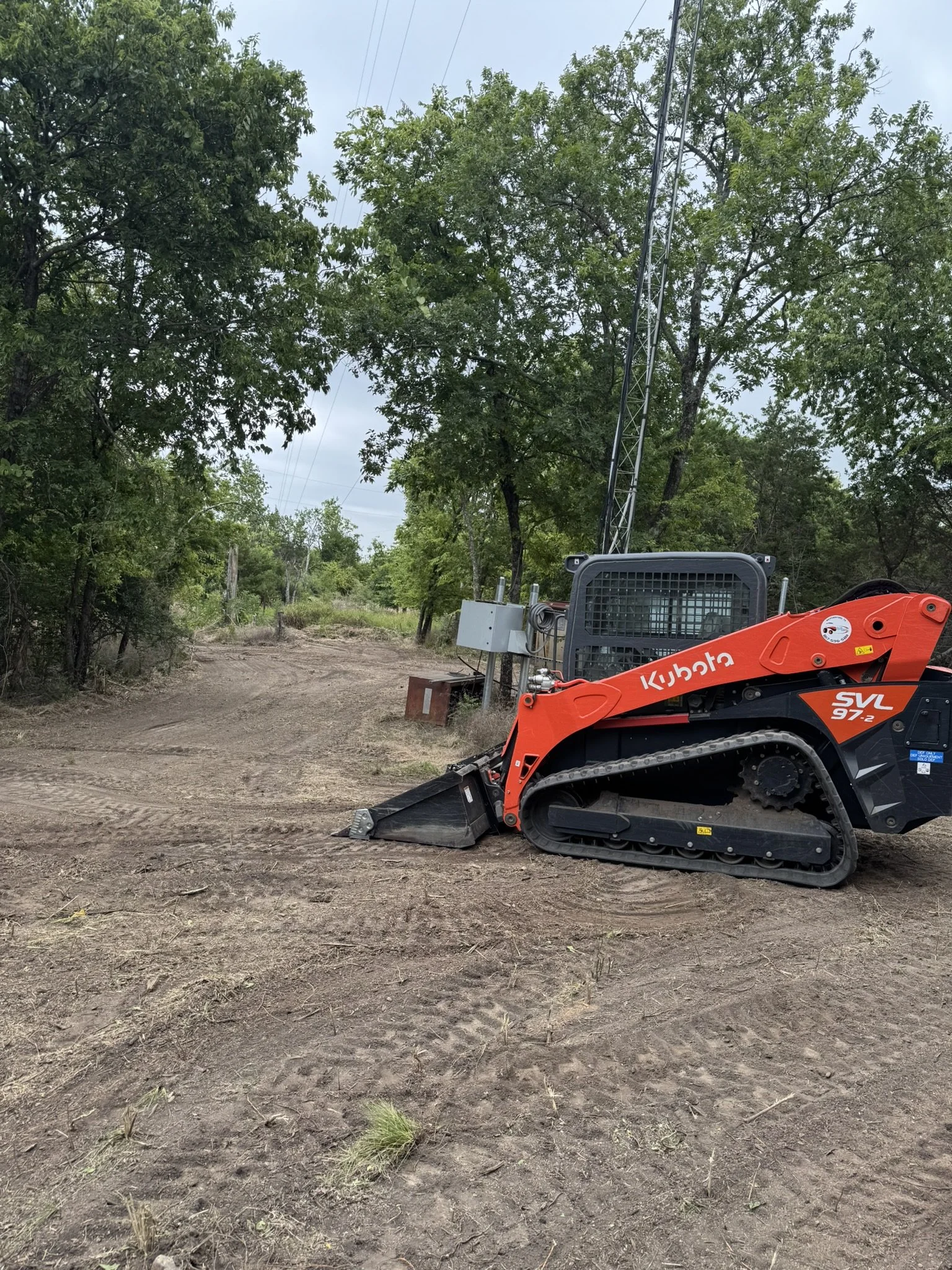 A construction site with a small orange Kubota compact excavator on a dirt surface, surrounded by green trees, electrical/wire infrastructure, and a cloudy sky.