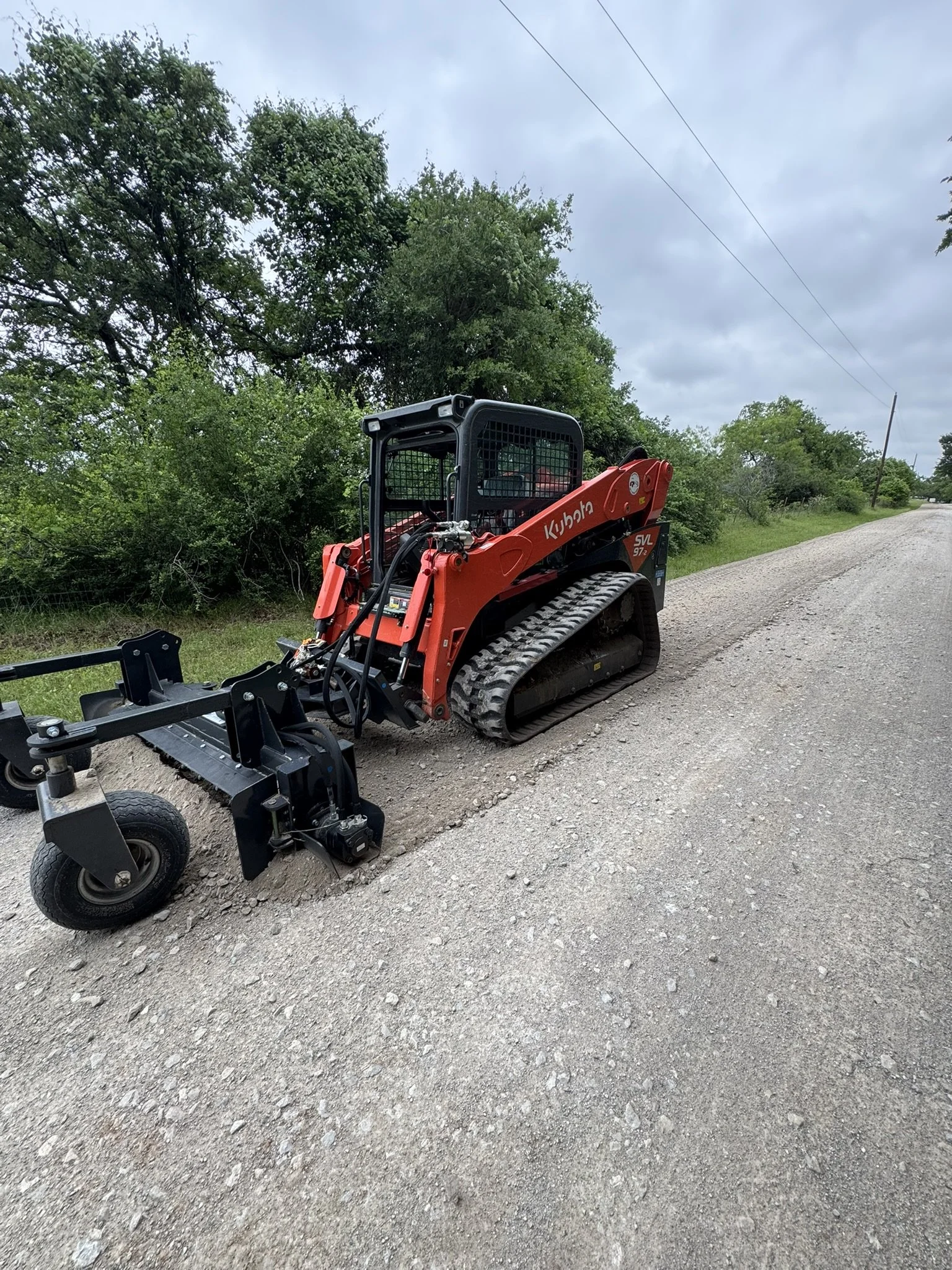 A small red Kubota mini excavator with caterpillar tracks parked on a gravel road next to green trees under a cloudy sky.