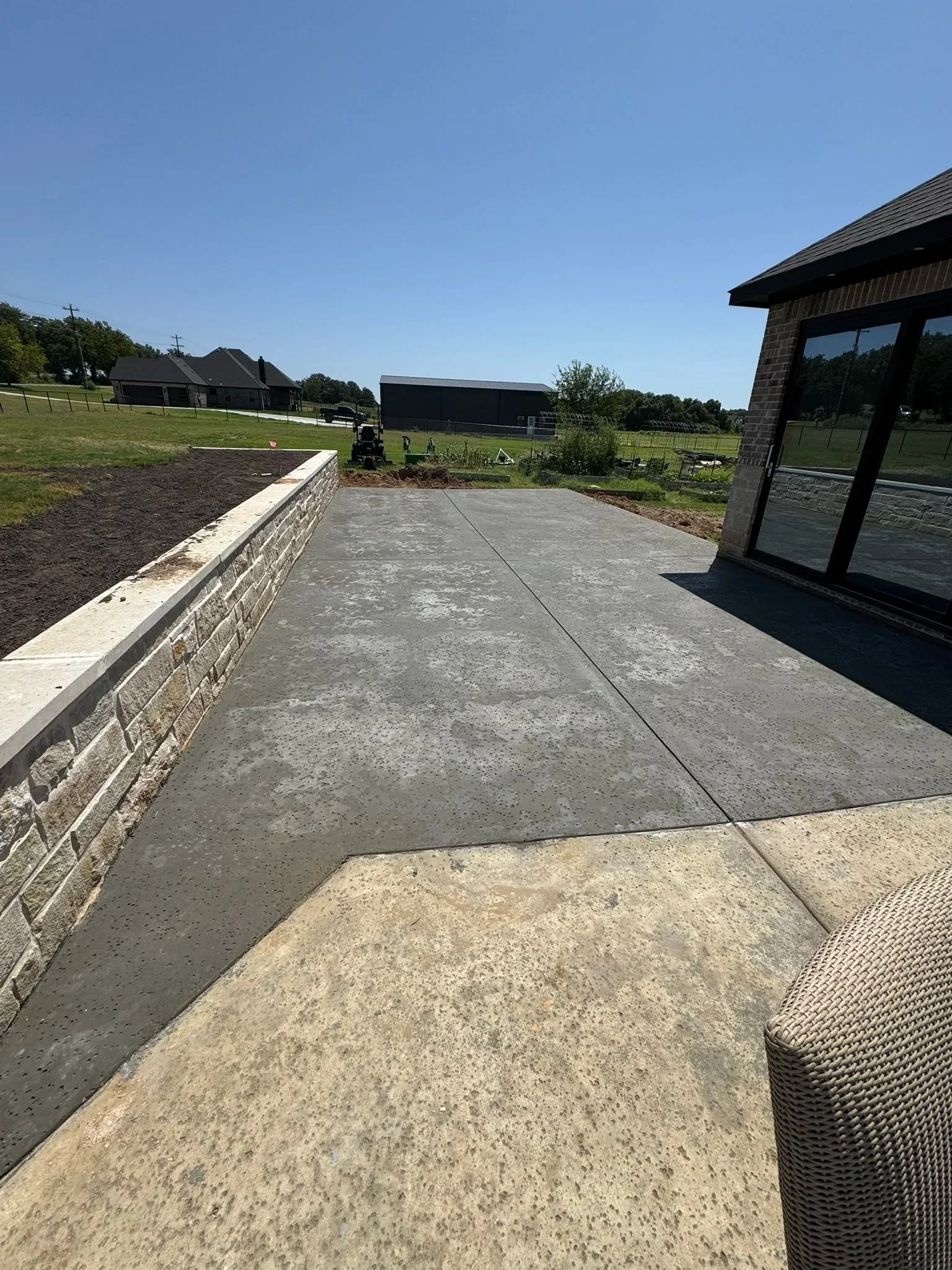 A newly concrete patio with a retaining wall on the left side, a wicker outdoor chair on the right, and a house with large windows on the right. In the background, there is a grassy yard, a farm building, and a clear blue sky.