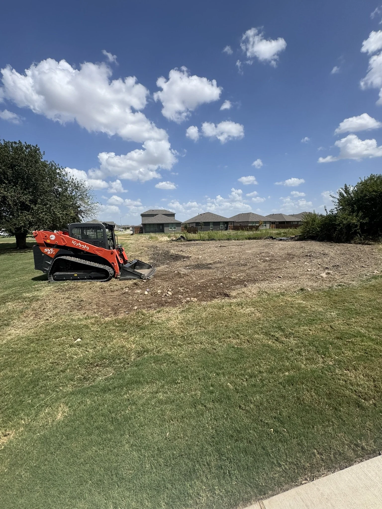 A construction site with a small orange Kubota SVL compact track loader on a patch of dirt, with green grass and residential houses in the background under a partly cloudy blue sky.