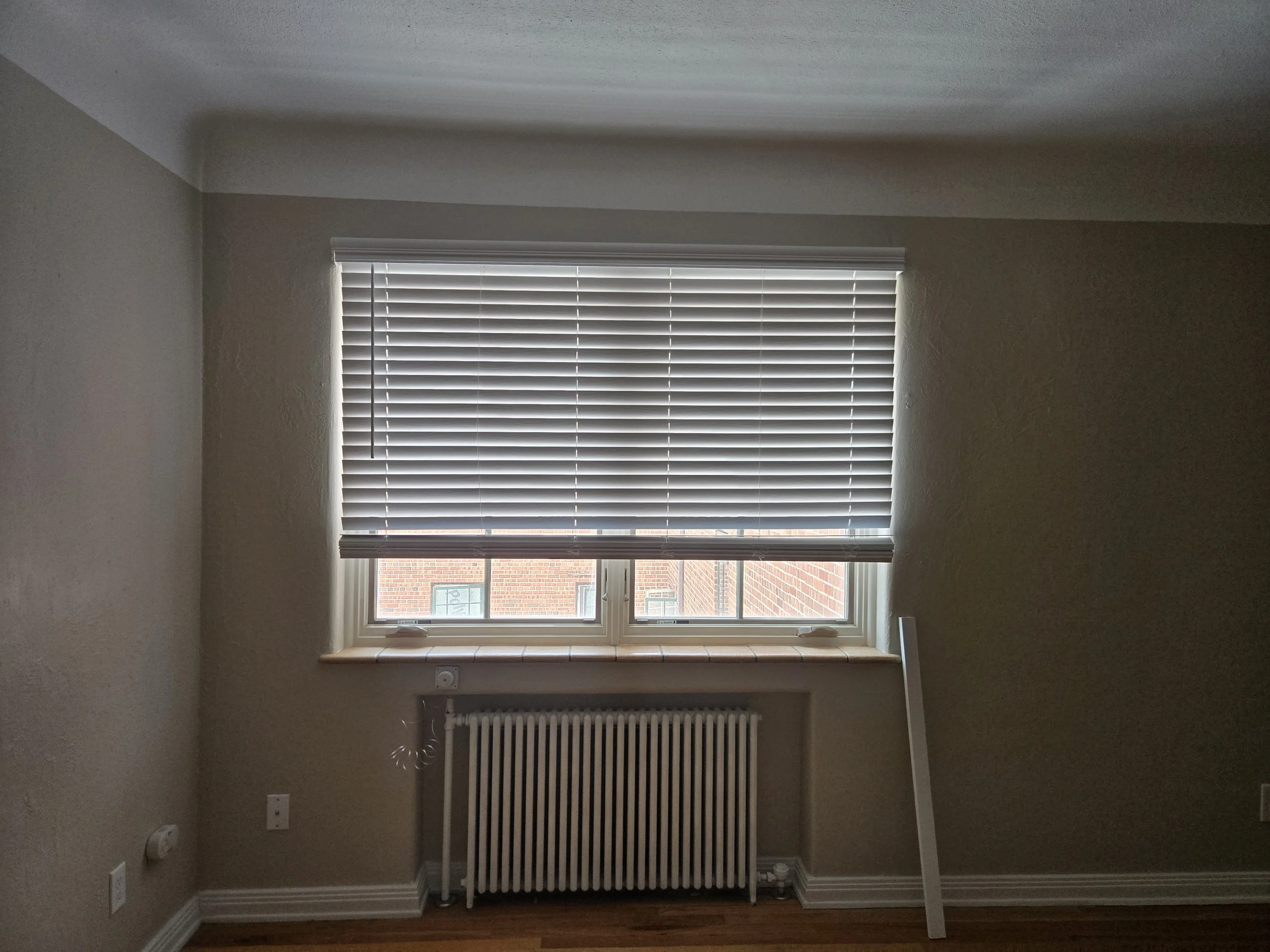 A room with a window covered by horizontal blinds, a radiator below the window, beige walls, and a hardwood floor.
