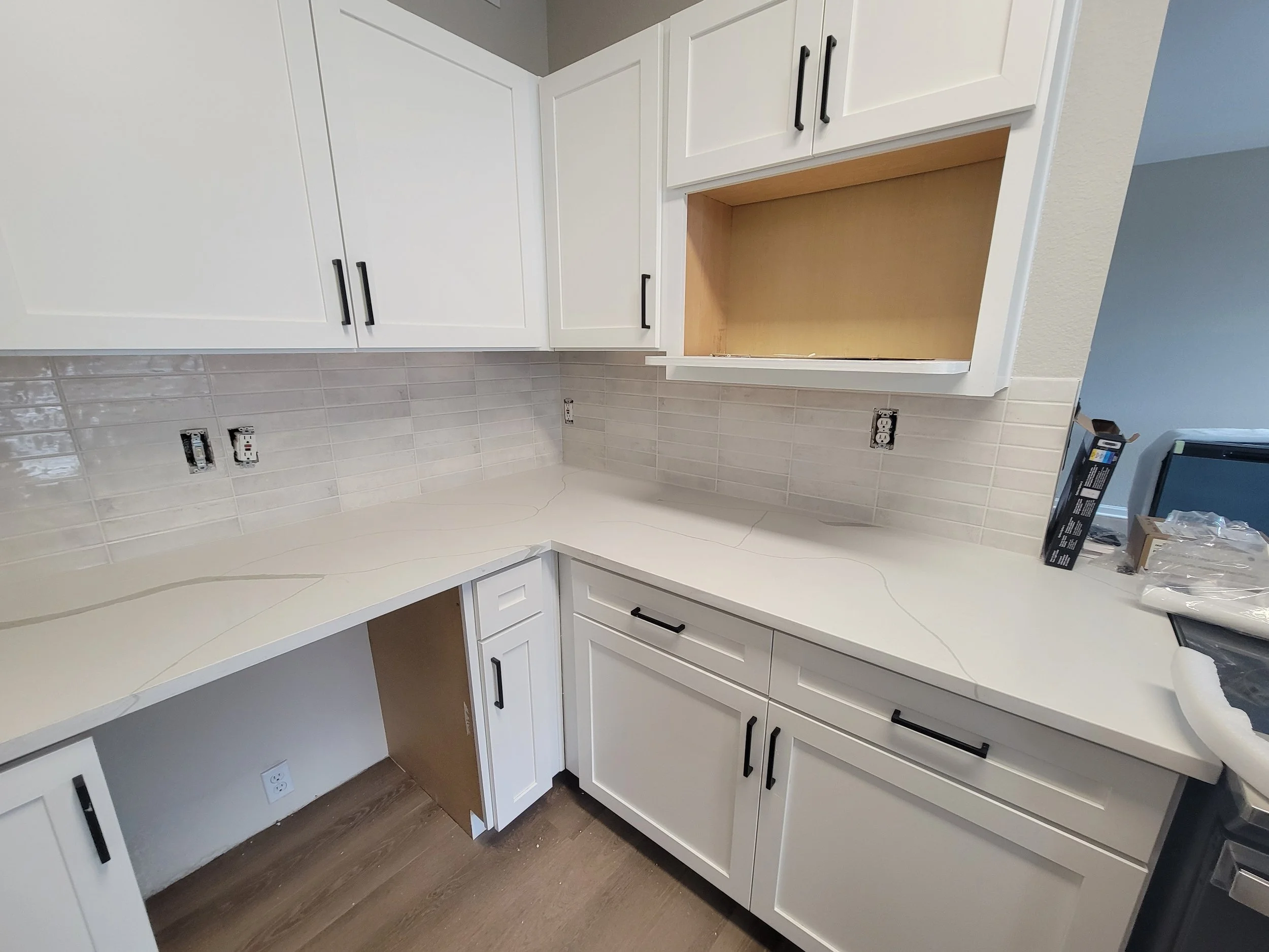 White kitchen cabinets with black handles, partially installed, with outlets and a countertop. An electrical outlet is visible on the wall below the cabinets.