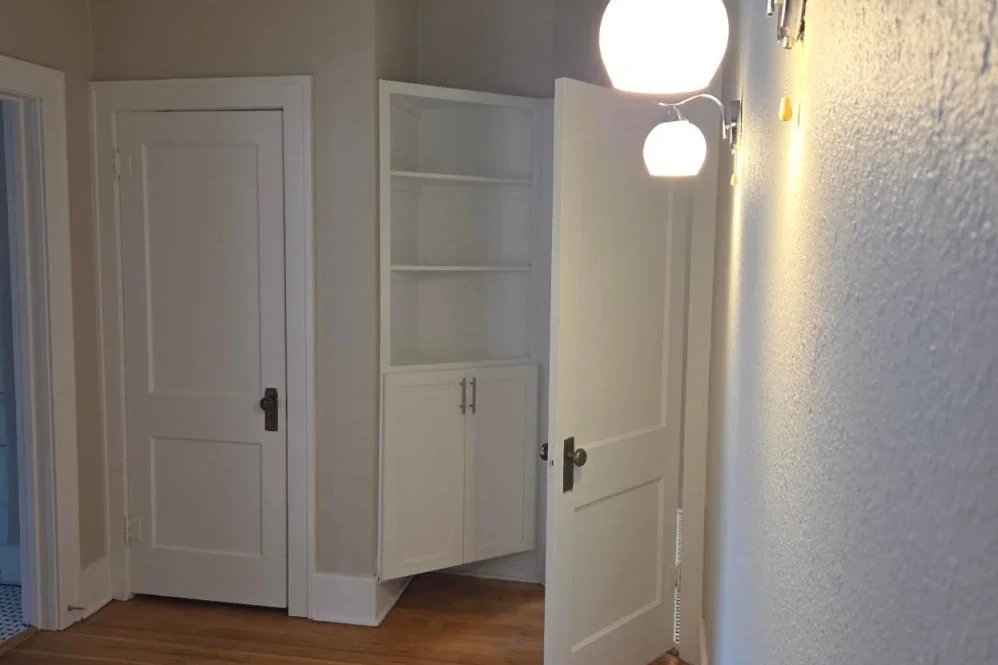 Open white door revealing a built-in white shelving cabinet in a room with beige walls and wooden floor, illuminated by two hanging round white lights.
