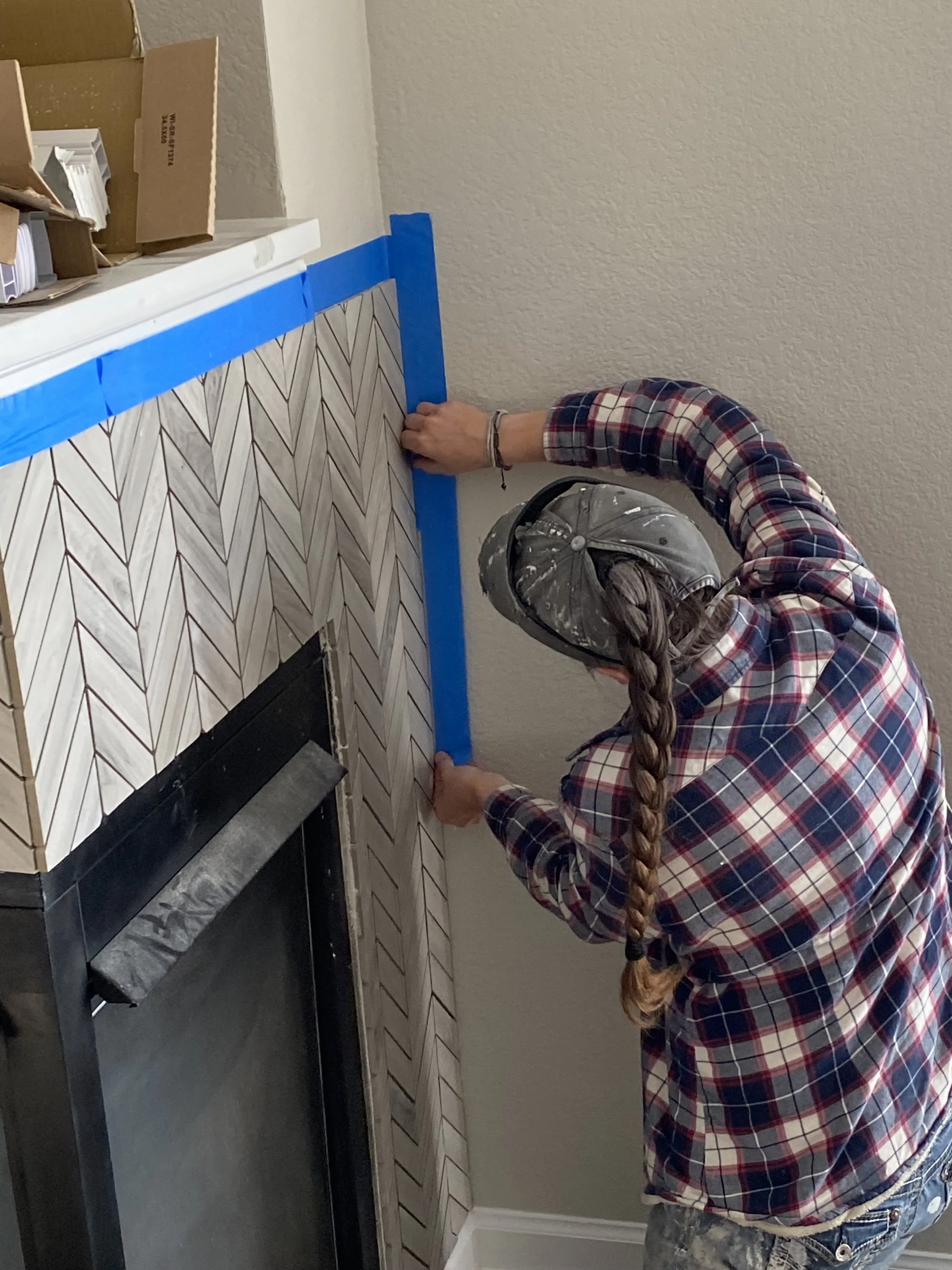 Person wearing a plaid shirt, denim jeans, and a cap, applying patterned tiles to a wall with blue painter’s tape along the edges for a home renovation project.