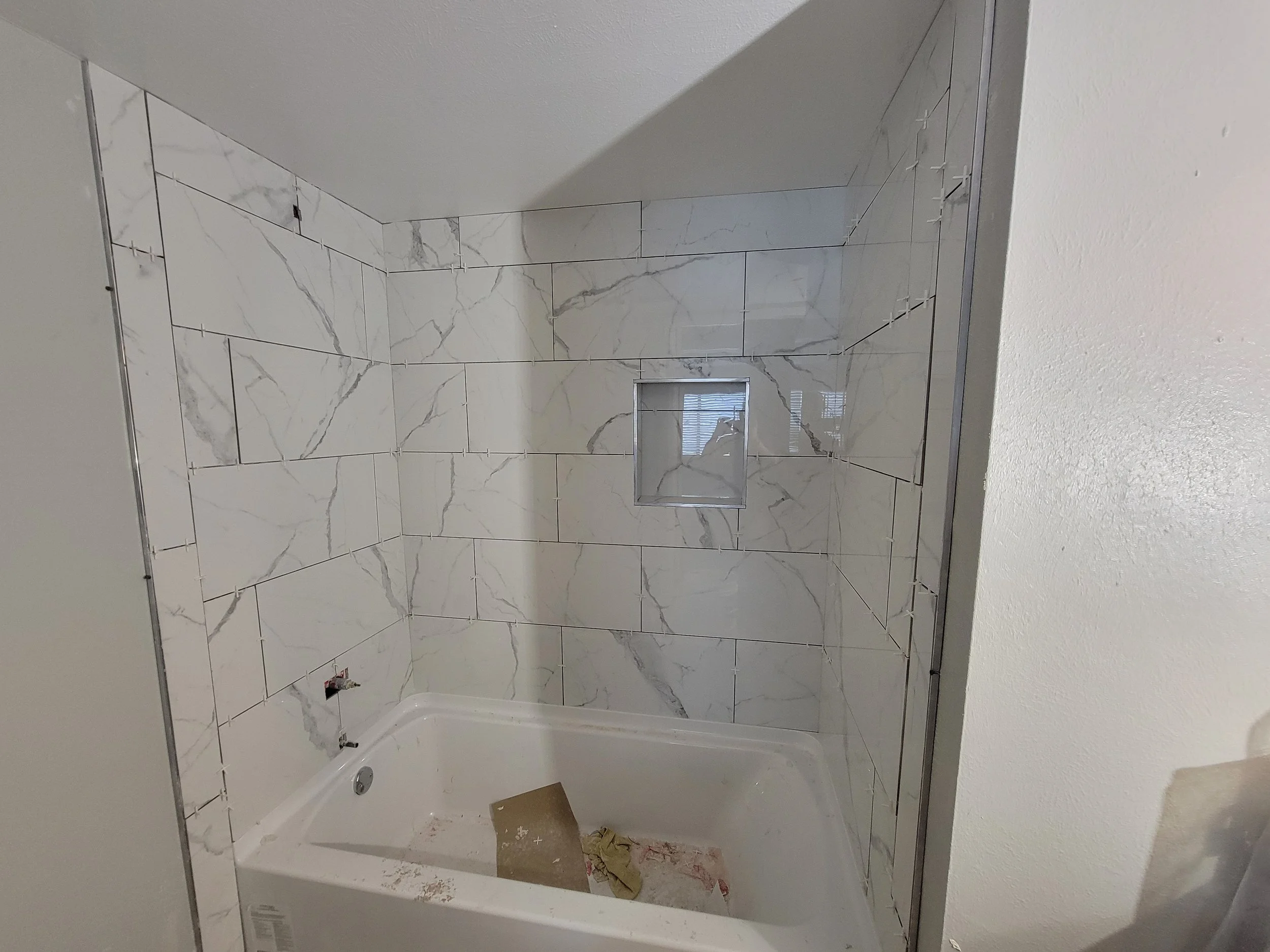 A bathroom shower area under construction with marble tiles partially installed, an open access panel, and construction debris on the floor.