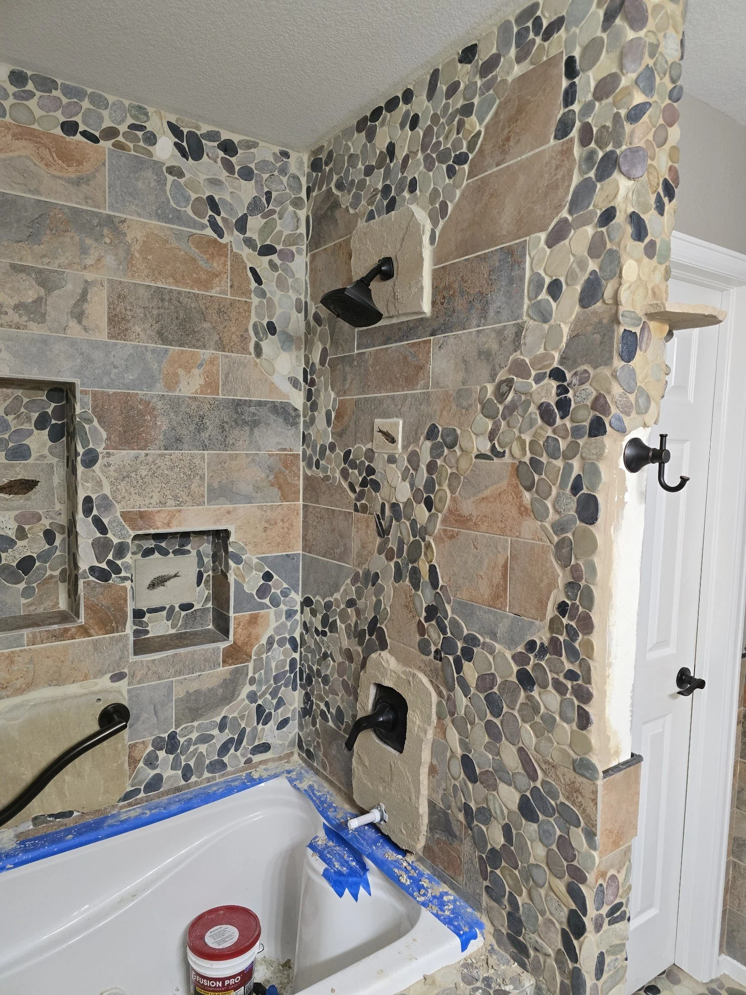 Bathroom wall with a decorative tile design featuring multicolored stones and rectangular tiles. Spray paint and tools are visible, indicating ongoing renovation.