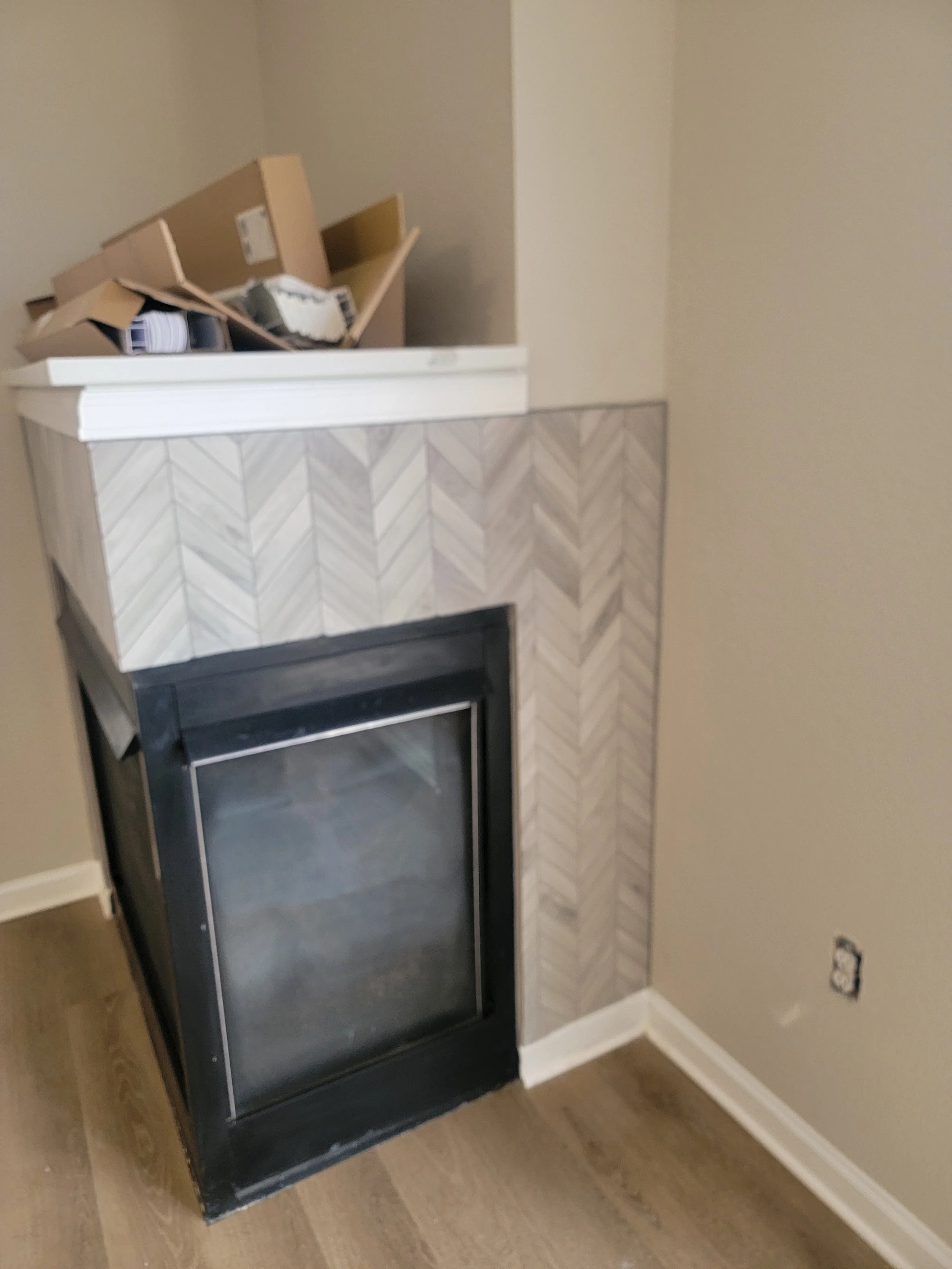 A fireplace with a patterned mantel, topped with an open cardboard box containing miscellaneous items, situated in a room with beige walls and wood flooring.