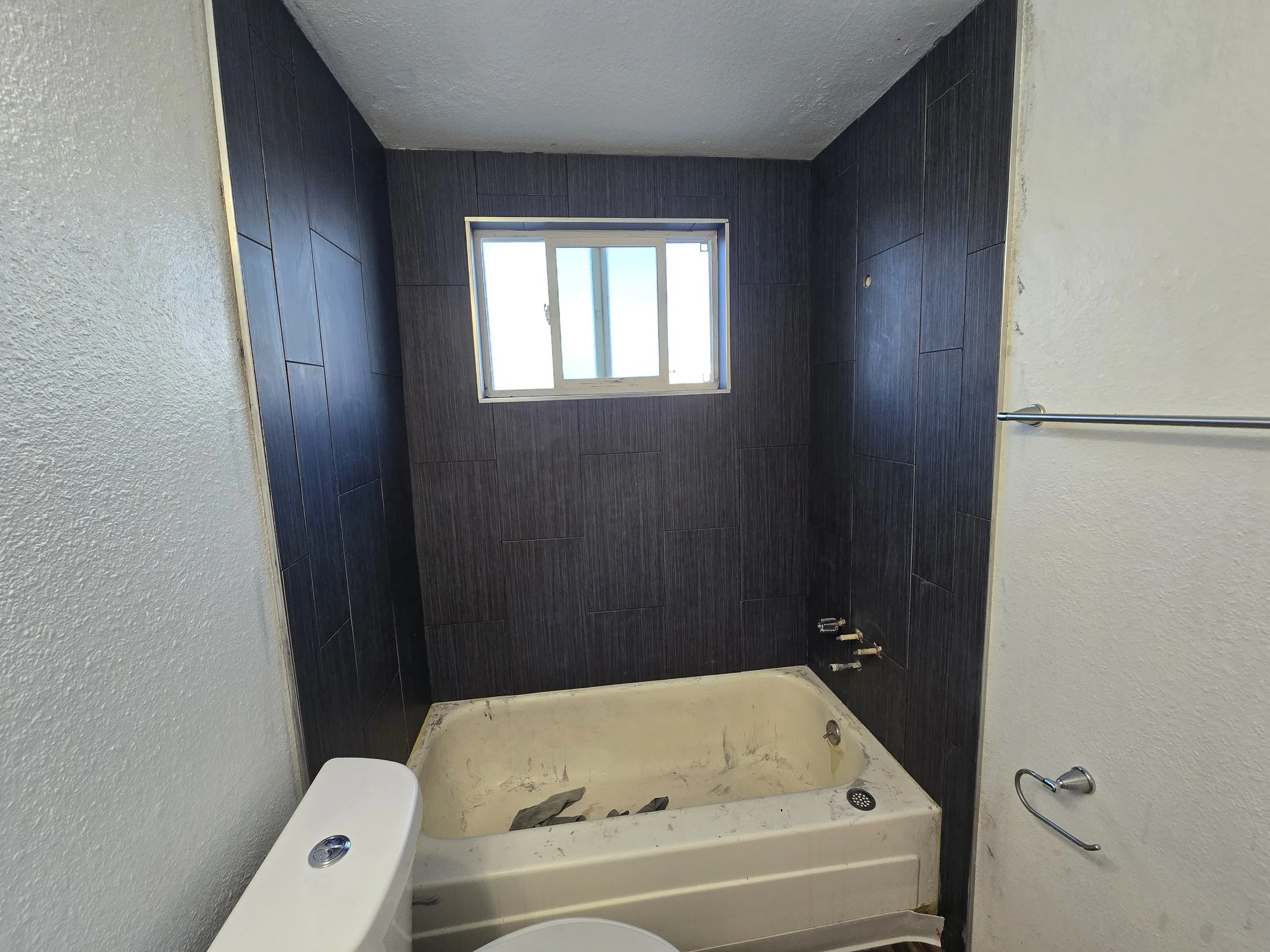 Bathroom under renovation with dark wall tiles, a window, a bathtub with construction debris and a toilet, partially installed.