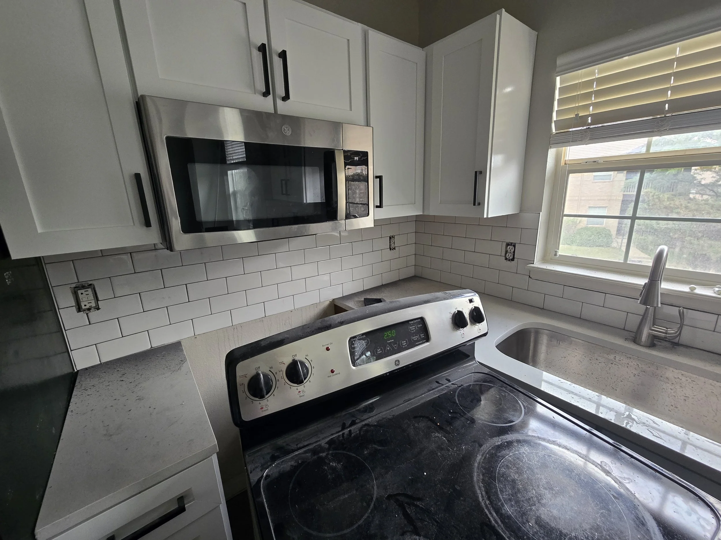A kitchen with white upper cabinets, a microwave above a stove, a window with blinds, and a sink with a faucet.