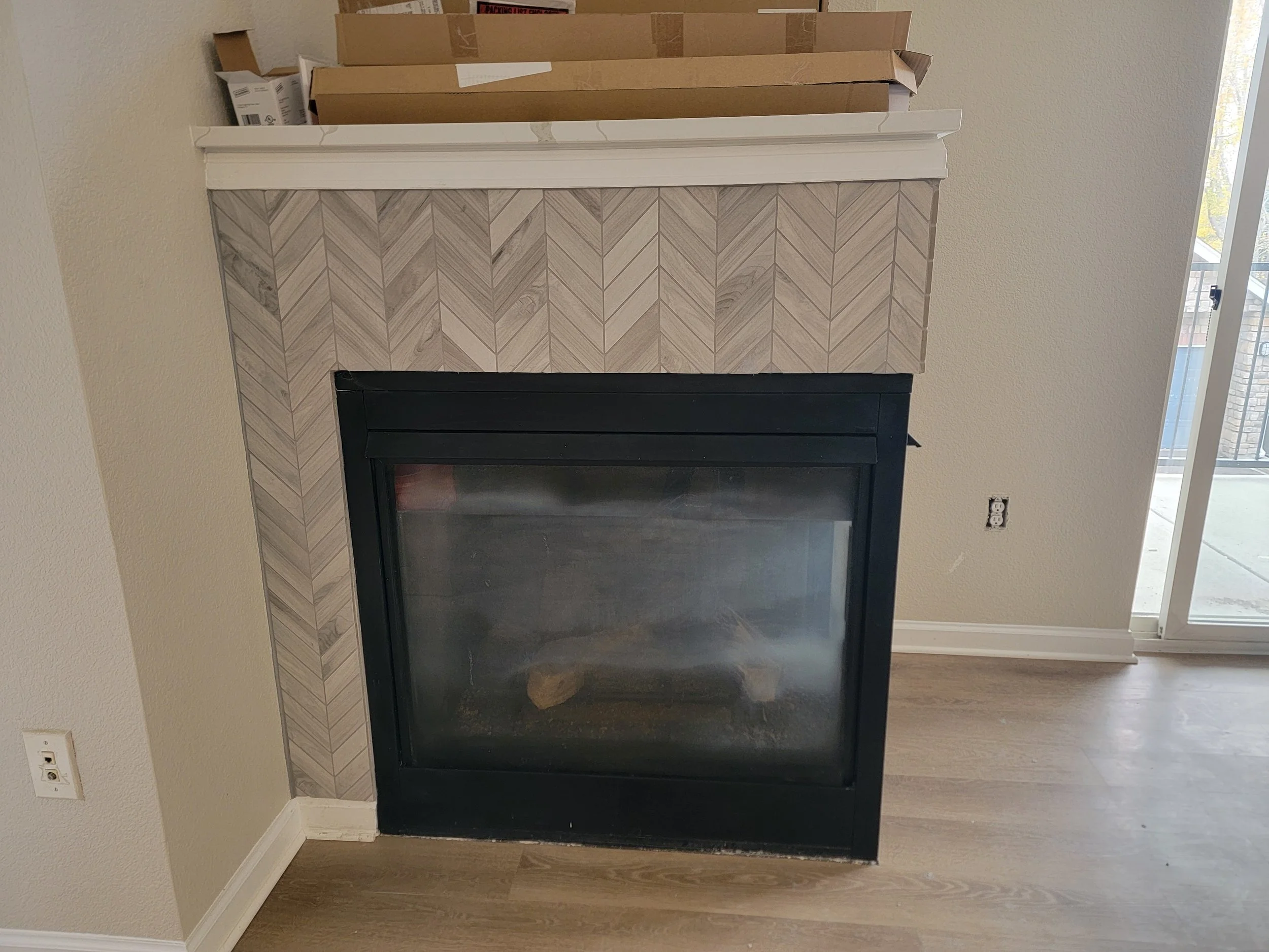 A living room fireplace with a beige and white patterned tile surround, lack of mantel, and set against a light beige wall. Several cardboard boxes are stacked above the fireplace on a wooden shelf, near a window with a view of a sunny outdoor area.