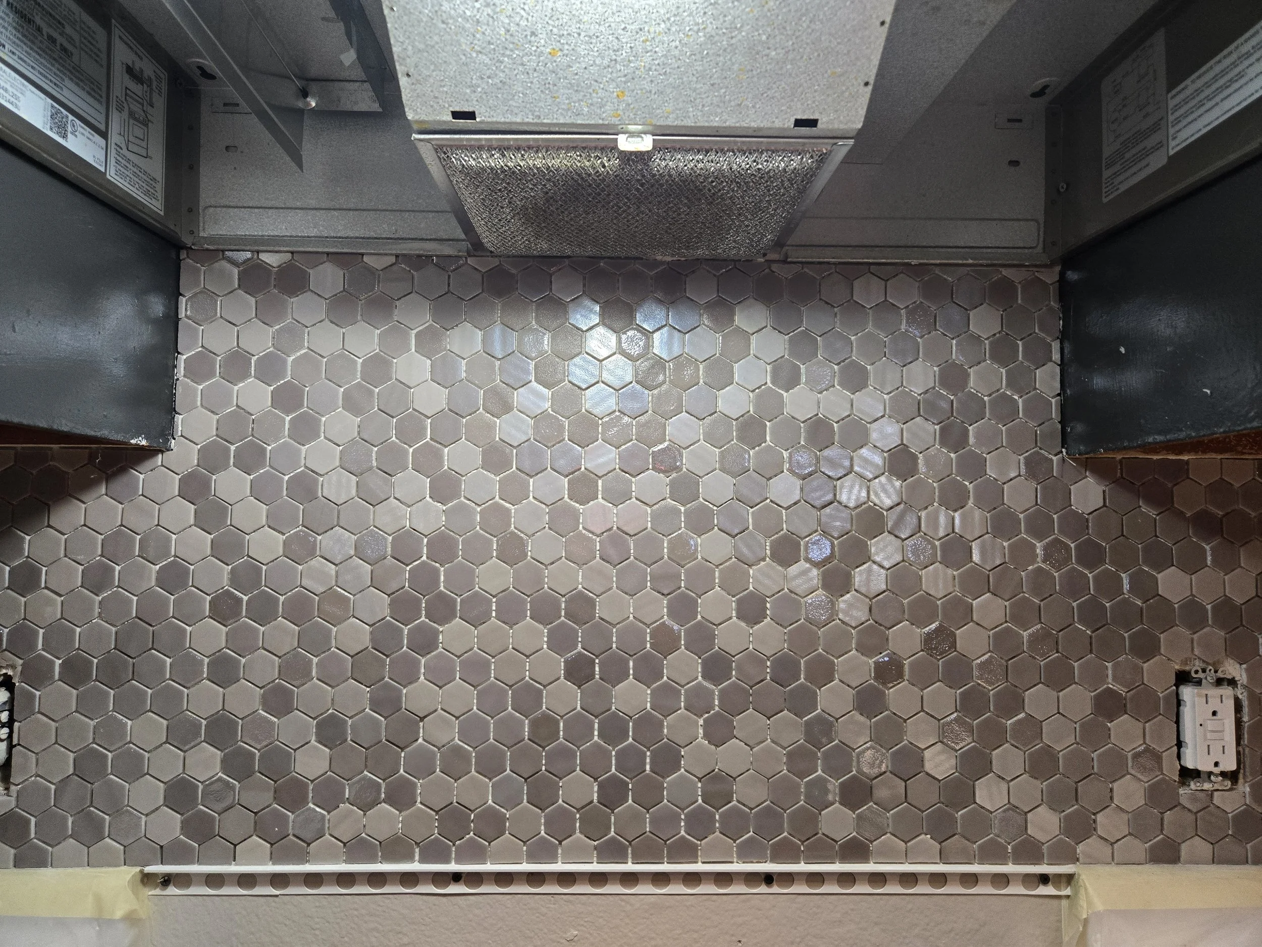 The image shows a section of a room with beige hexagonal tile flooring, electrical outlets, and a metallic vent on the ceiling. There is a strip of tape along the bottom edge of the wall.